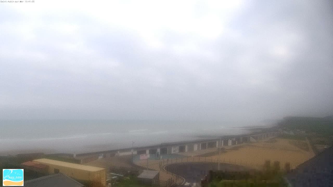 A wide coastal view shows a choppy sea washing onto a sandy beach alongside a long promenade and buildings, all beneath a heavily overcast and grey sky.