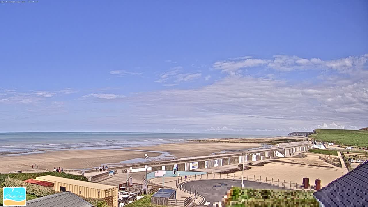 A wide coastal view shows a choppy sea washing onto a sandy beach alongside a long promenade and buildings, all beneath a heavily overcast and grey sky.