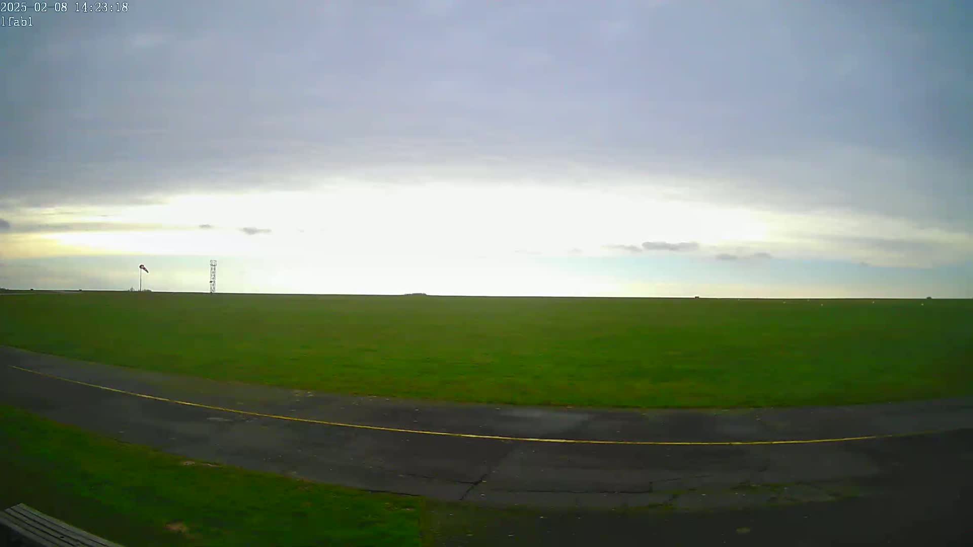 A large green field stretches to the horizon under a mostly cloudy sky, with a paved road curving in the foreground.
