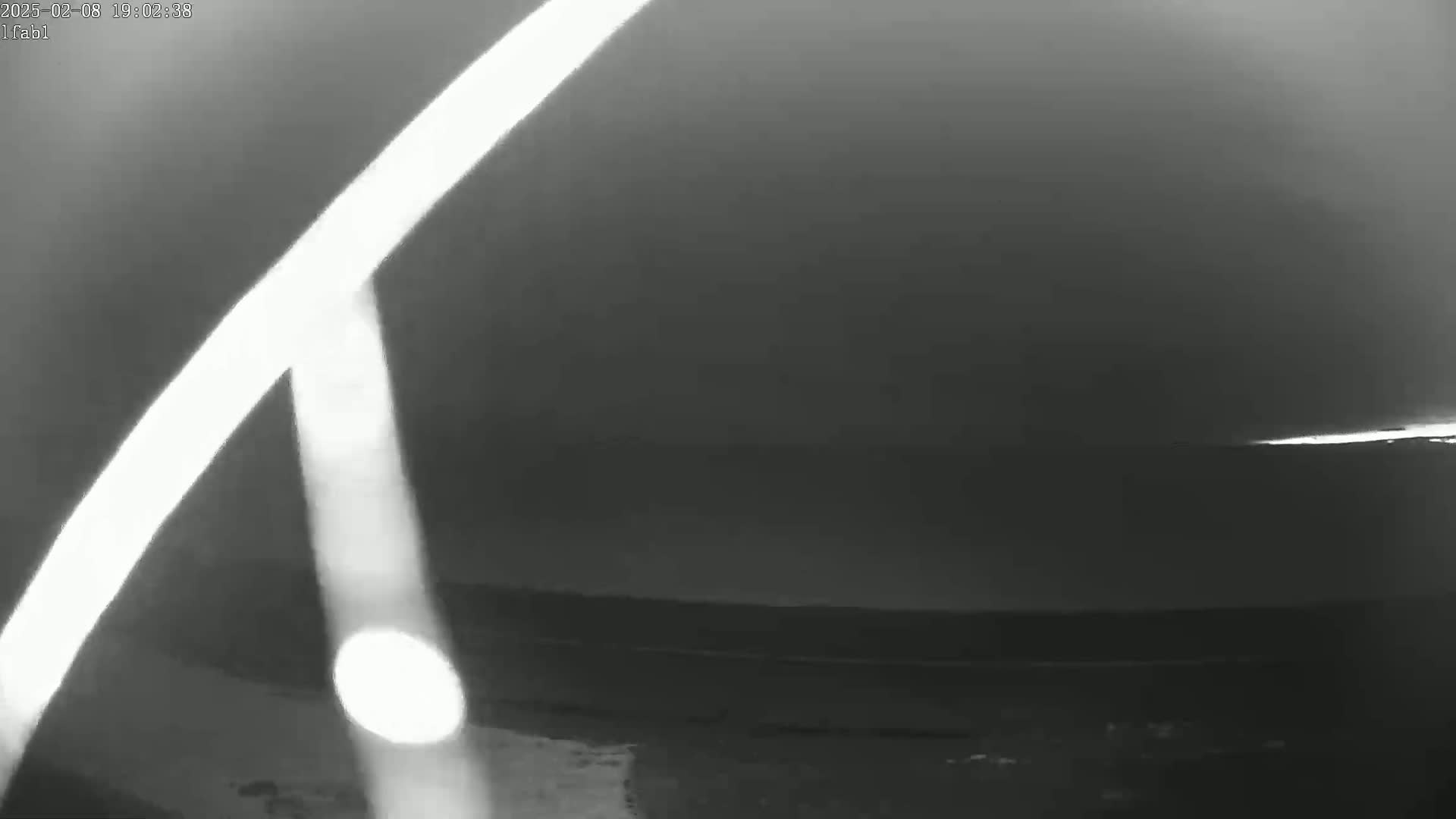 A low-light grayscale image shows bright streaks of light possibly from headlights against a dark, overcast sky above a flat horizon line.
