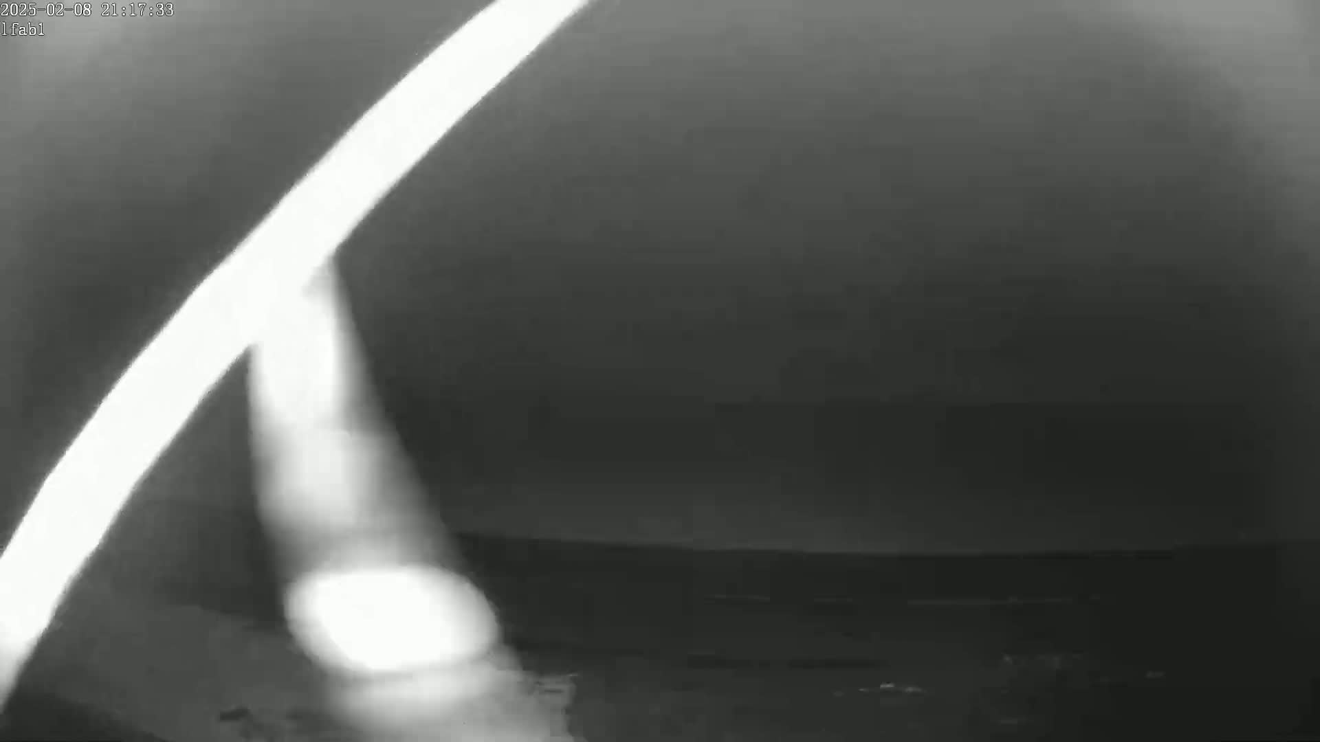 A blurry, low-light nighttime image shows a bright light source, possibly a streetlight, in the bottom left corner with a bright streak that appears to be a light reflection or glare in the upper left corner, against a dark, mostly featureless background.
