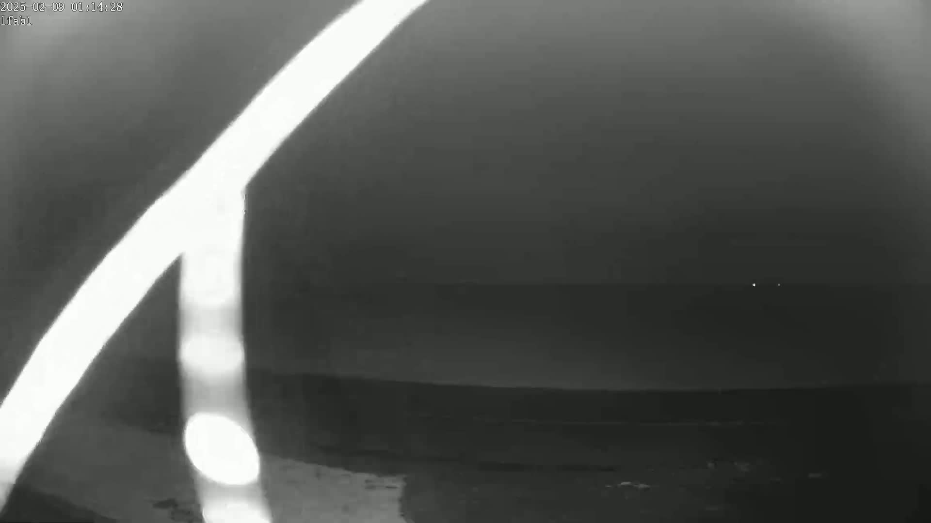 A nighttime, low-light image shows a blurry, bright white light source in the foreground with a dark body of water and faint lights visible on the horizon.