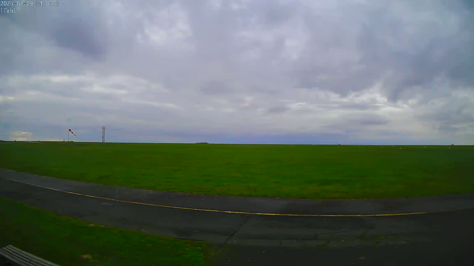 A grassy field under an overcast sky features a paved road curving into the foreground, with a windsock and a tall structure visible in the mid-ground.