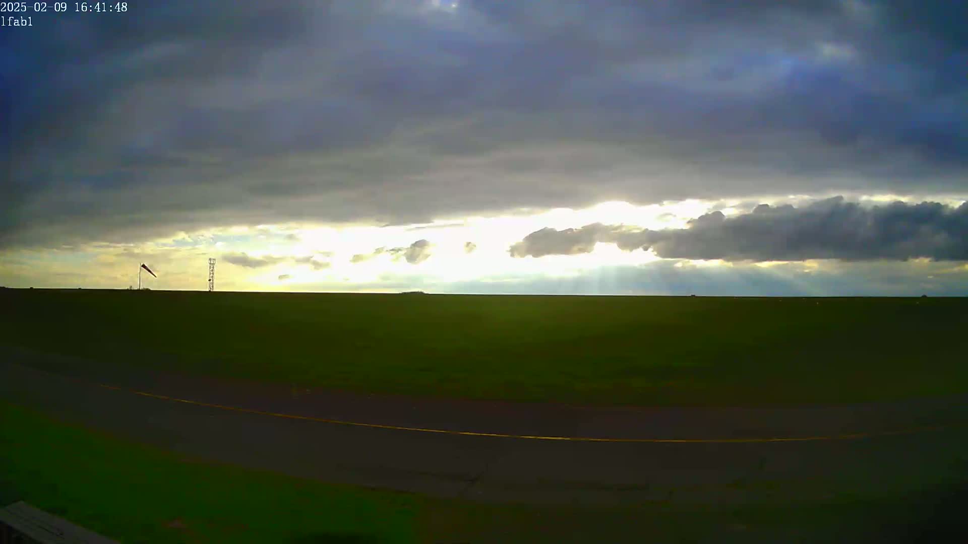 A flat green field with a paved road stretches out under a partly cloudy sky, where sunlight breaks through the clouds.