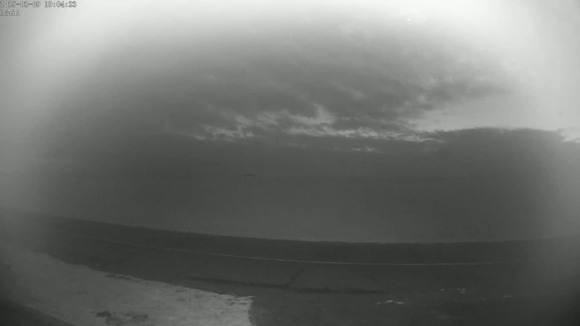 A grayscale image shows a mostly cloudy sky over a flat, dark landscape at dusk.