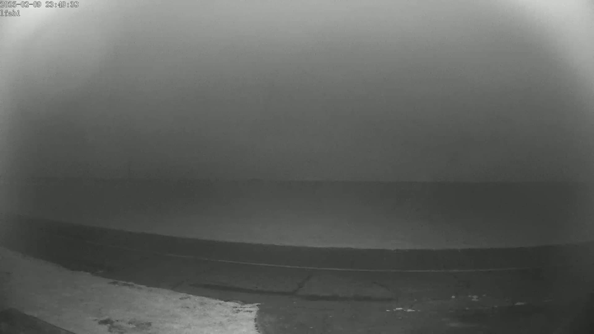 A low-visibility, foggy scene shows a dark, flat expanse of land or water near a road or shoreline at night.