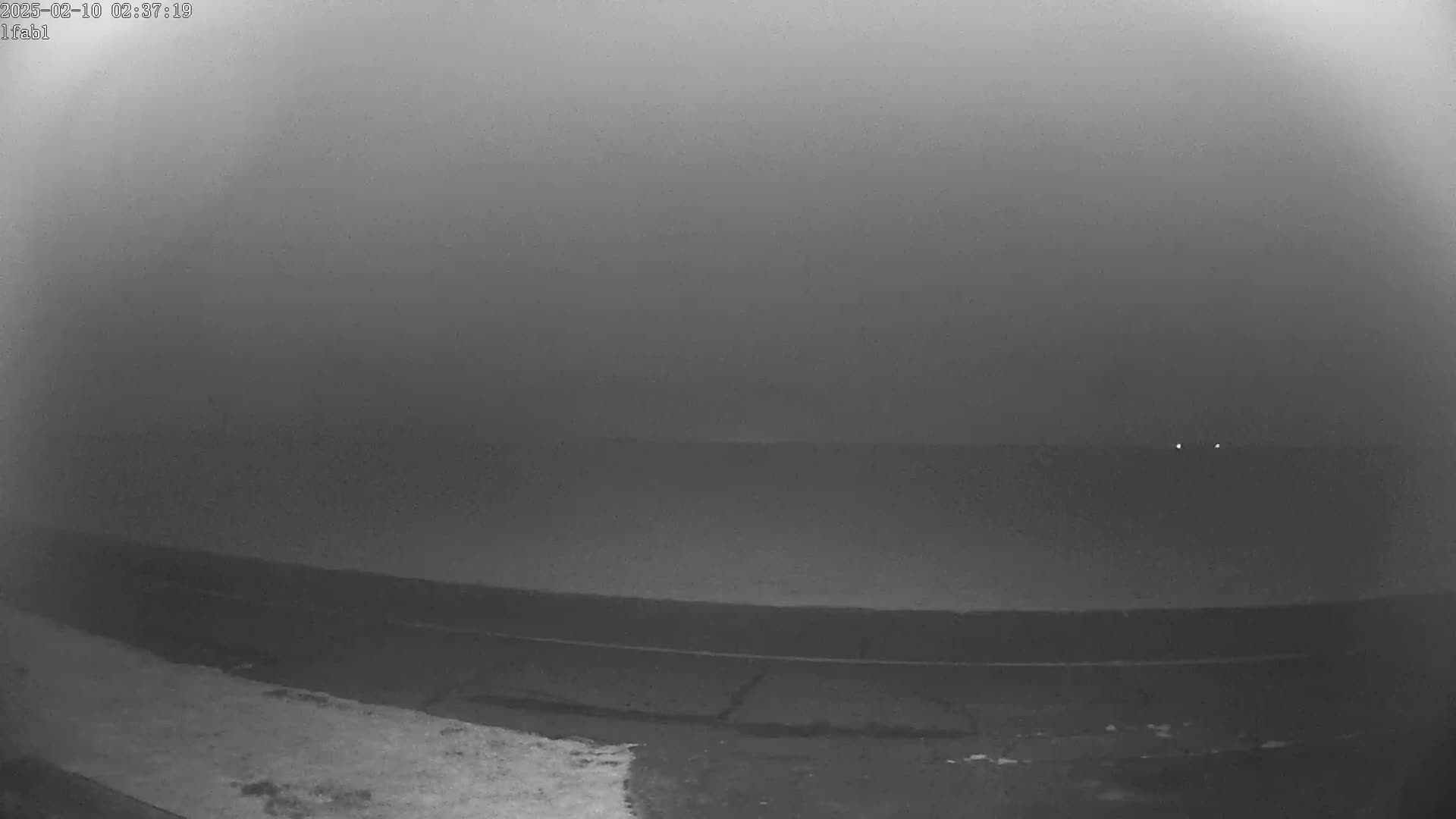 A foggy, dark outdoor scene shows a flat expanse of land leading to a body of water with two small lights visible in the distance.