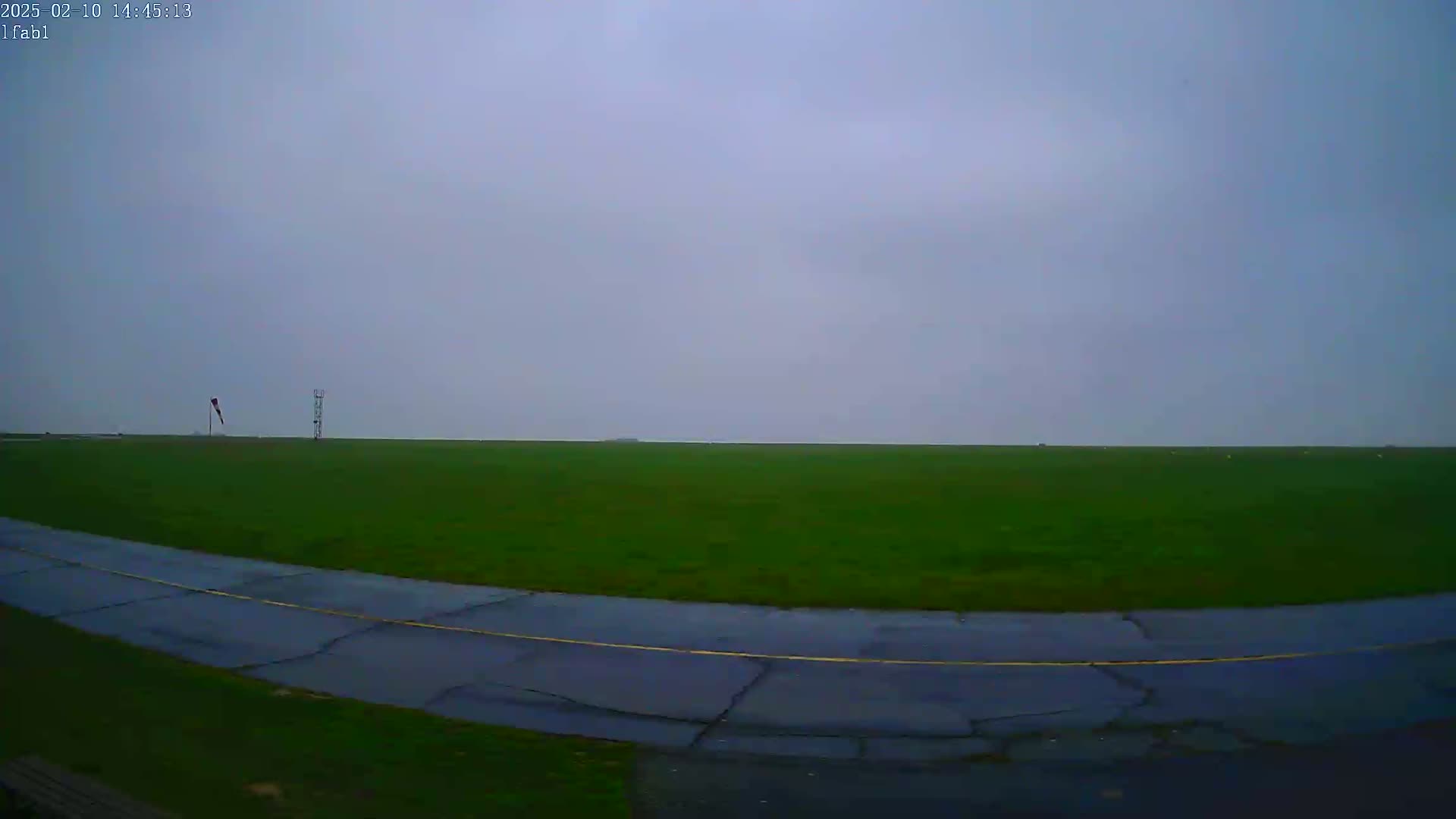 A wet, dark gray day reveals a grassy airfield with a curved taxiway and a windsock.