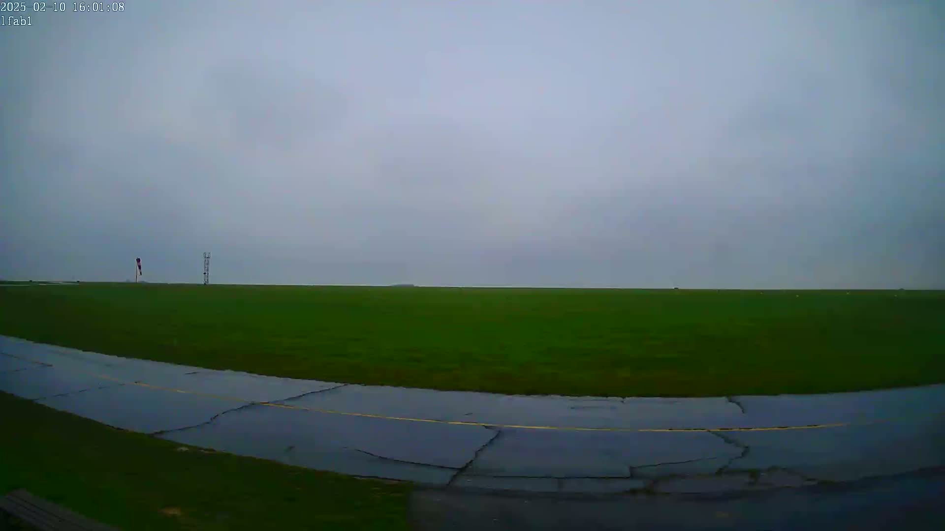 A wet, paved taxiway curves through a large, grassy field under an overcast sky.