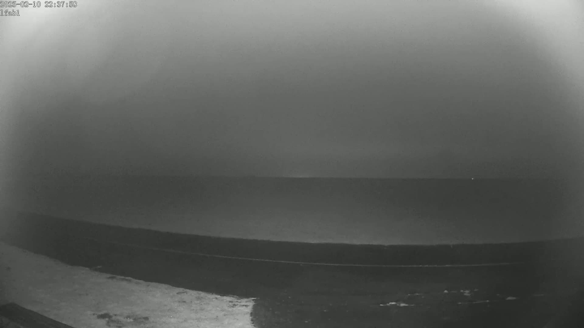 A dark, foggy scene shows a body of water meeting a dark shoreline under a hazy sky.