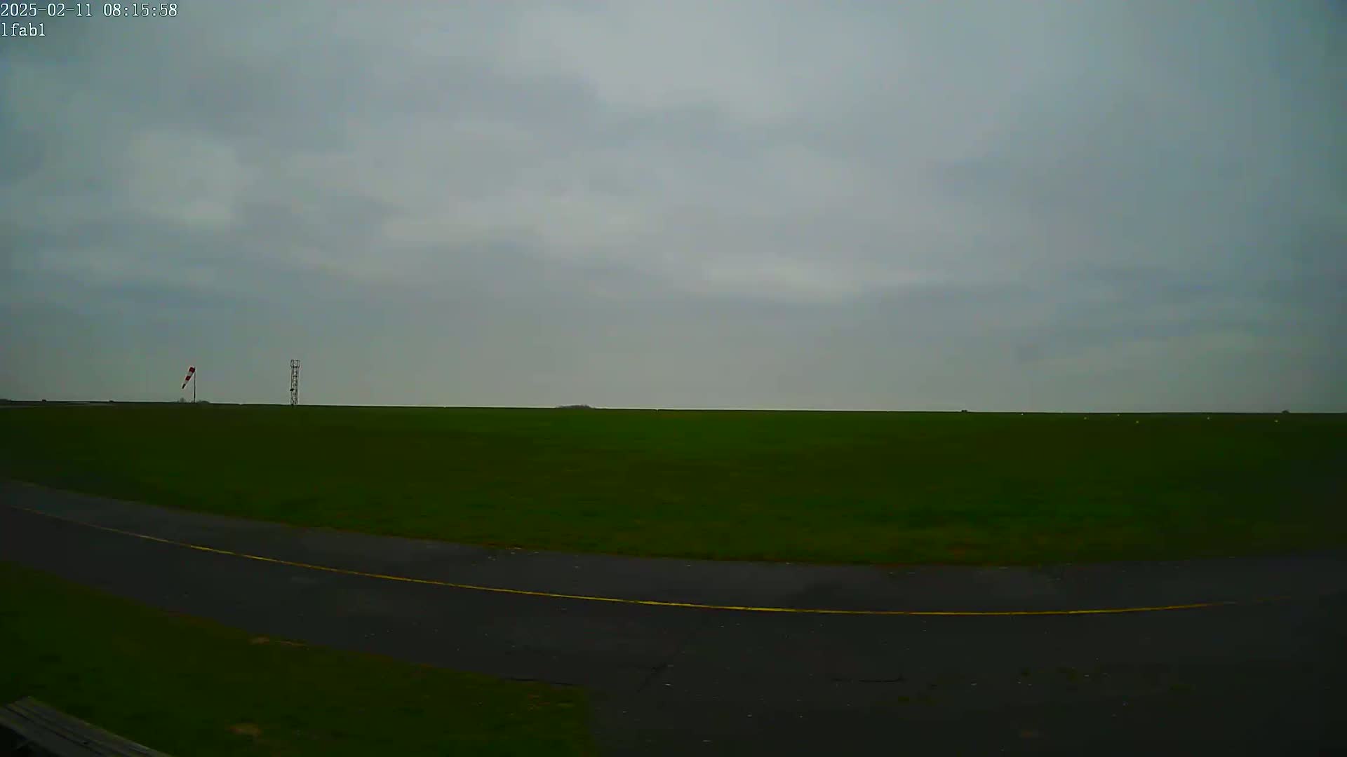 A flat, green field stretches to a horizon under an overcast sky, with a paved road curving in the foreground and a windsock and tall structure visible in the distance.