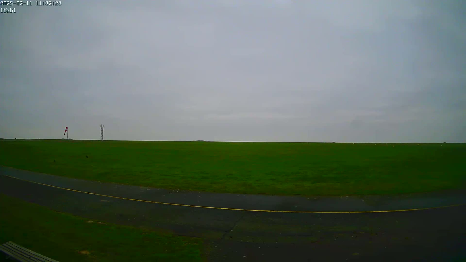 A wide, flat expanse of green grass under an overcast sky stretches to the horizon, with a paved road curving in the foreground and a small windsock and tower visible in the mid-ground.