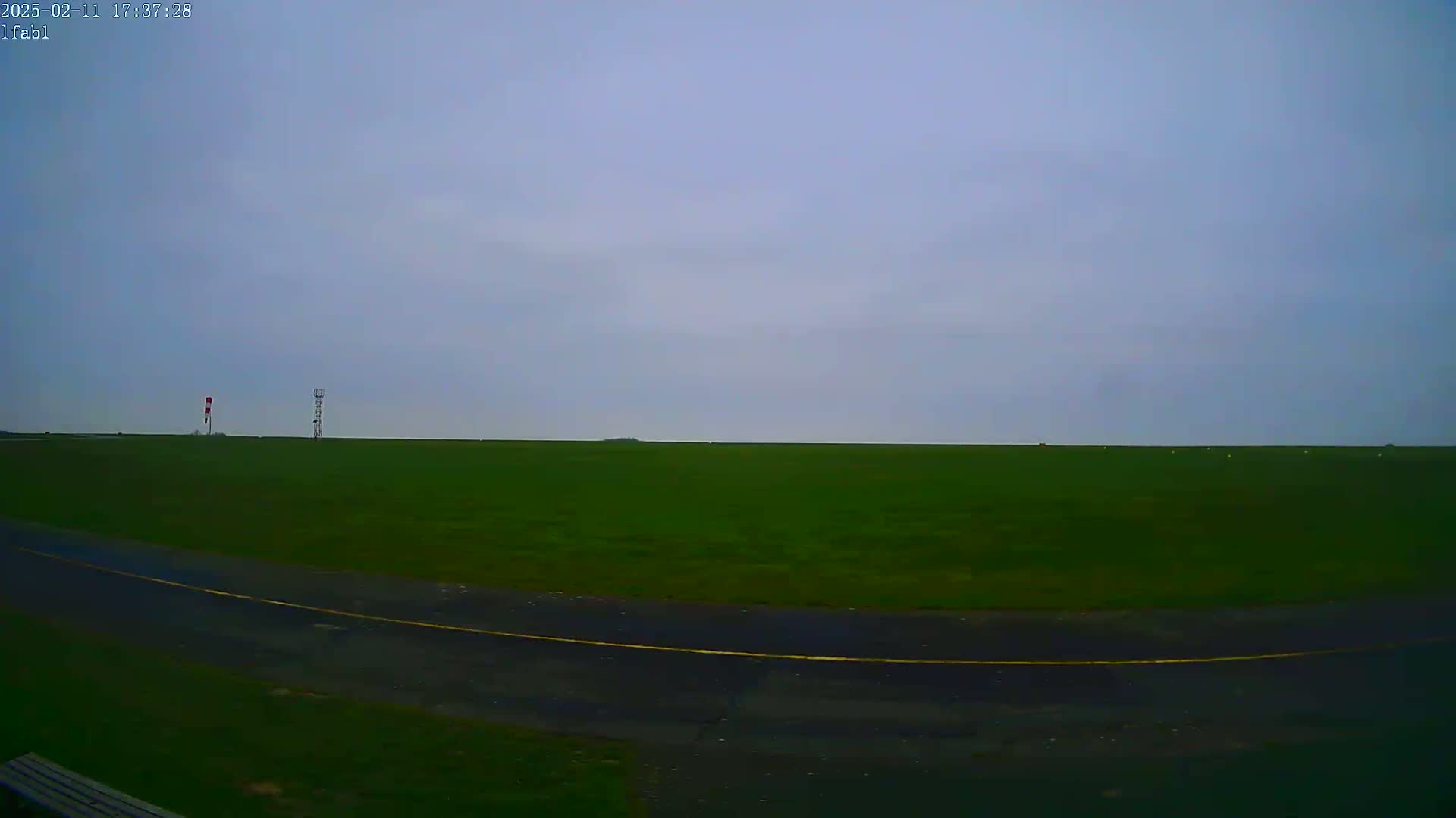 A flat, green field stretches to the horizon under a cloudy, overcast sky, with a small paved area and two tall structures in the distance.
