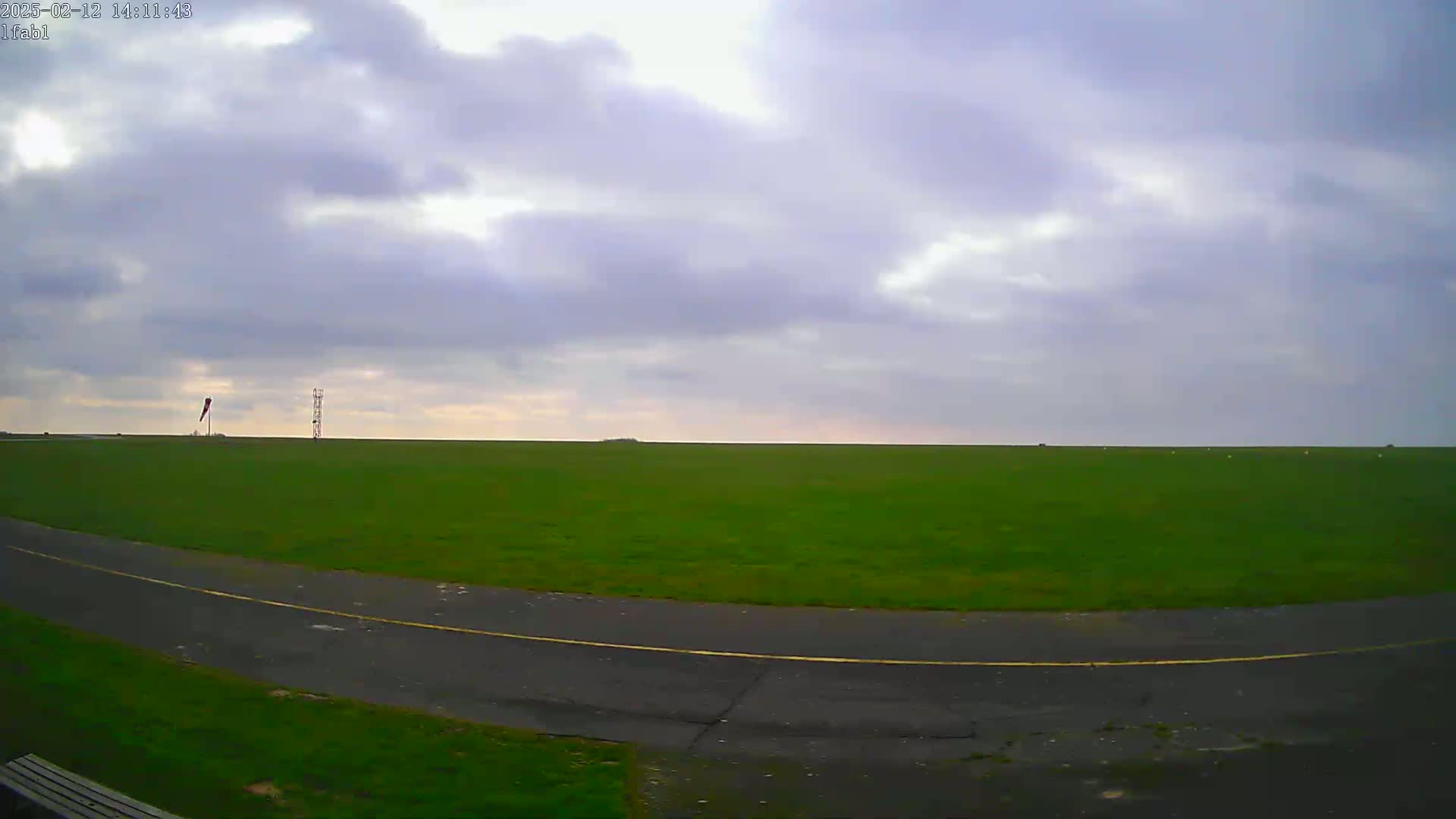 A paved path curves through a large, flat expanse of green grass under a mostly cloudy sky.