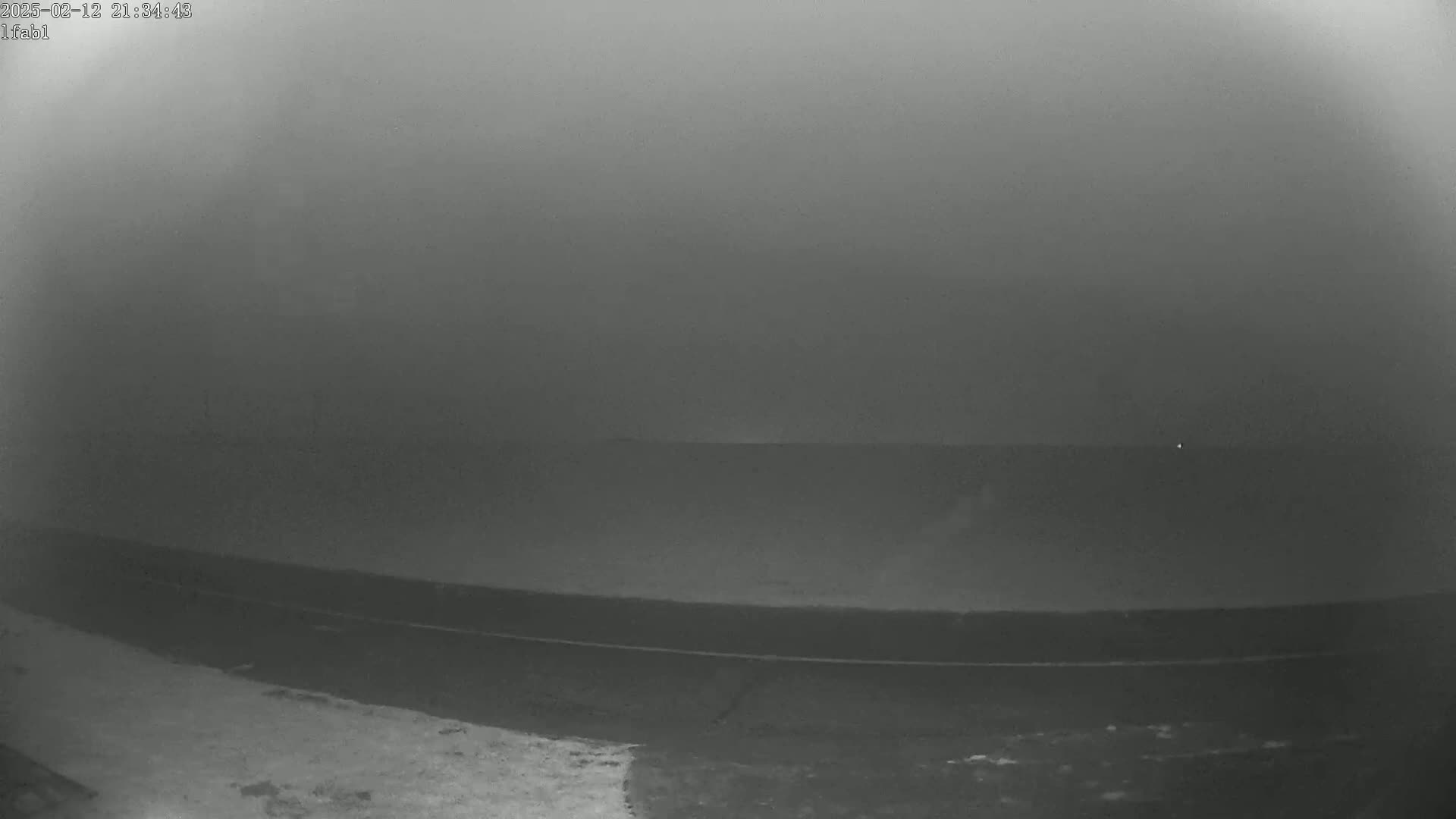 A foggy, dark scene shows a flat expanse of land and a dark body of water, possibly a lake or ocean, under a gray sky.