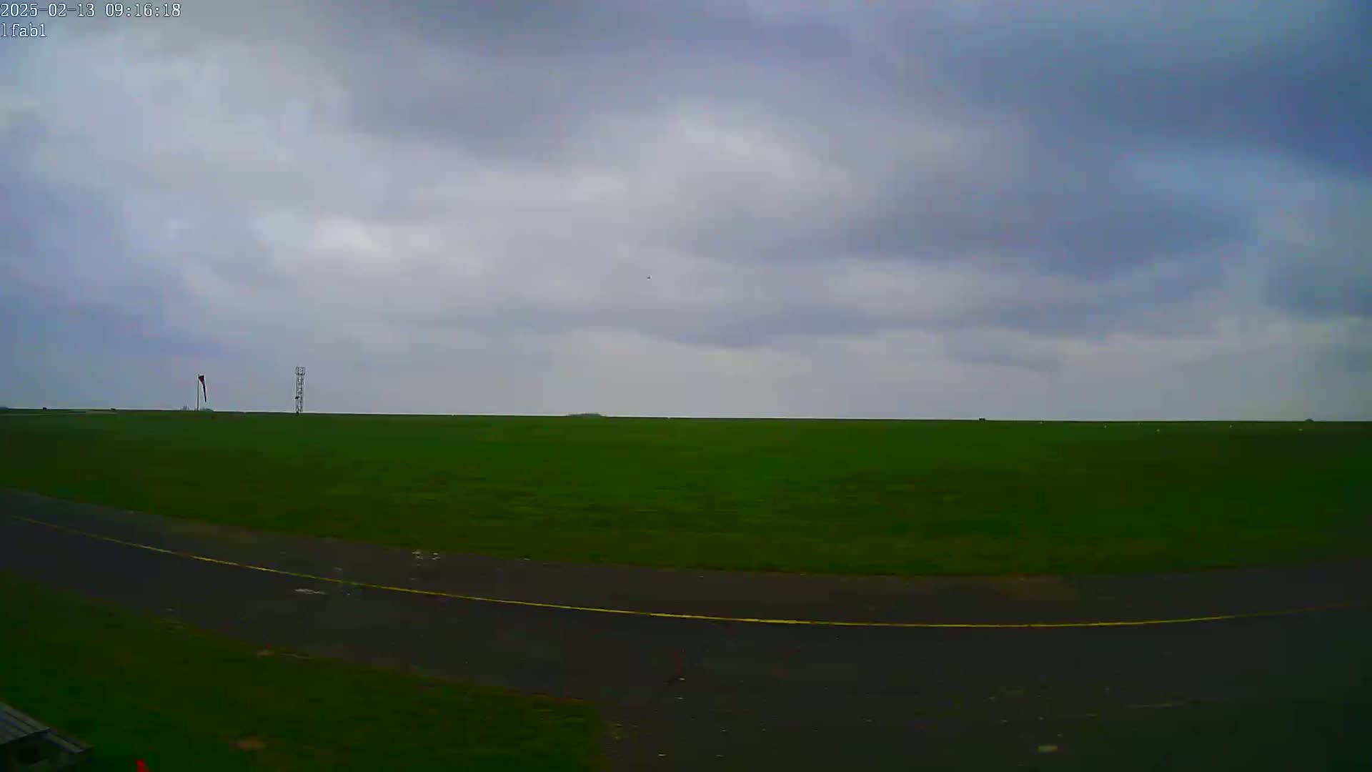 A wide, flat expanse of green grass under a cloudy sky, with a paved area and a small windsock visible in the distance.