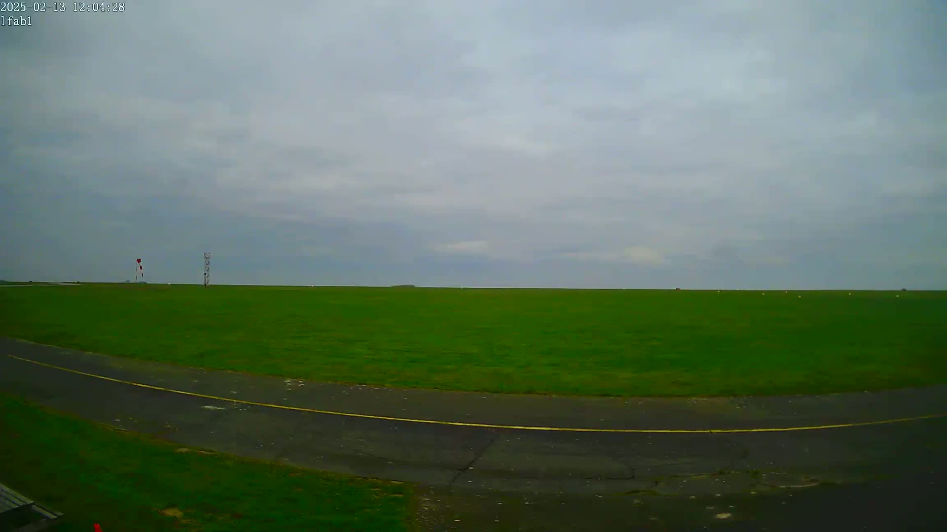 A wide, grassy field under an overcast sky is bordered by a paved road curving into the foreground.