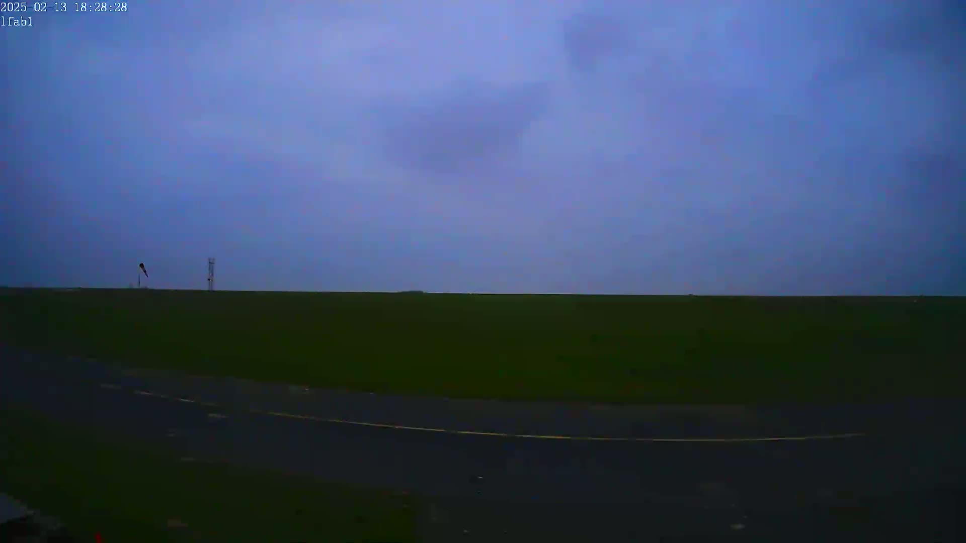 A flat, dark green field stretches to a dusky blue sky under overcast conditions, with a road and two tall structures visible in the distance.
