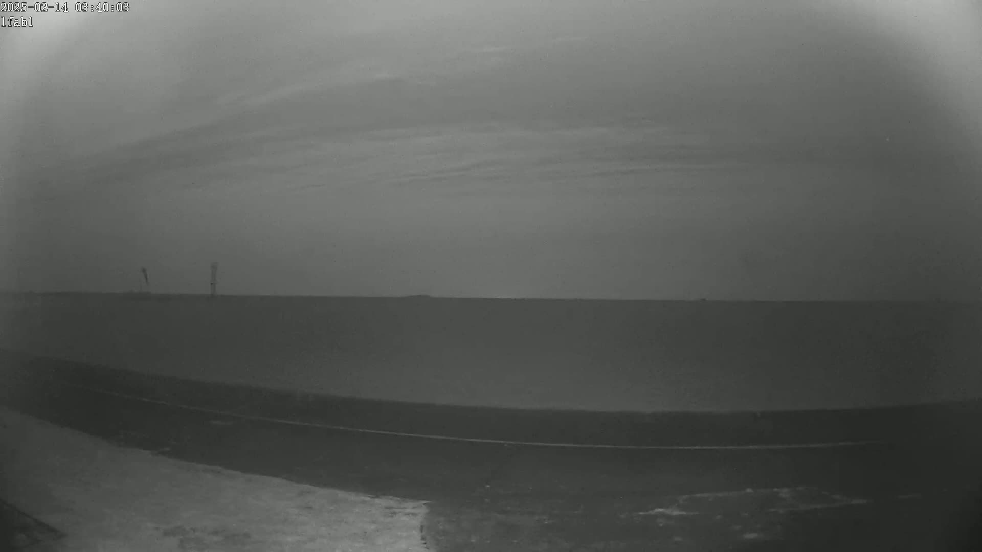 A low-light, overcast outdoor scene shows a flat, dark landscape with two distant, thin structures and a dark road or path in the foreground.