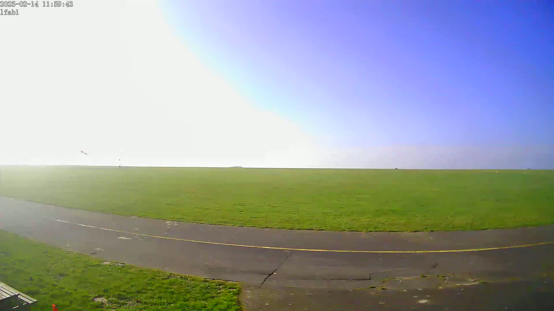 A paved path curves through a large grassy field under a mostly clear, bright blue sky.