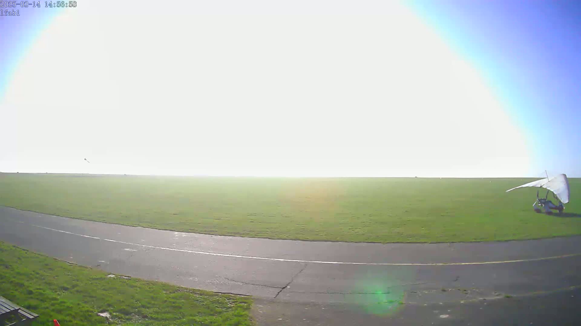 A hang glider sits on a grassy airfield with a paved runway under a bright, mostly sunny sky.