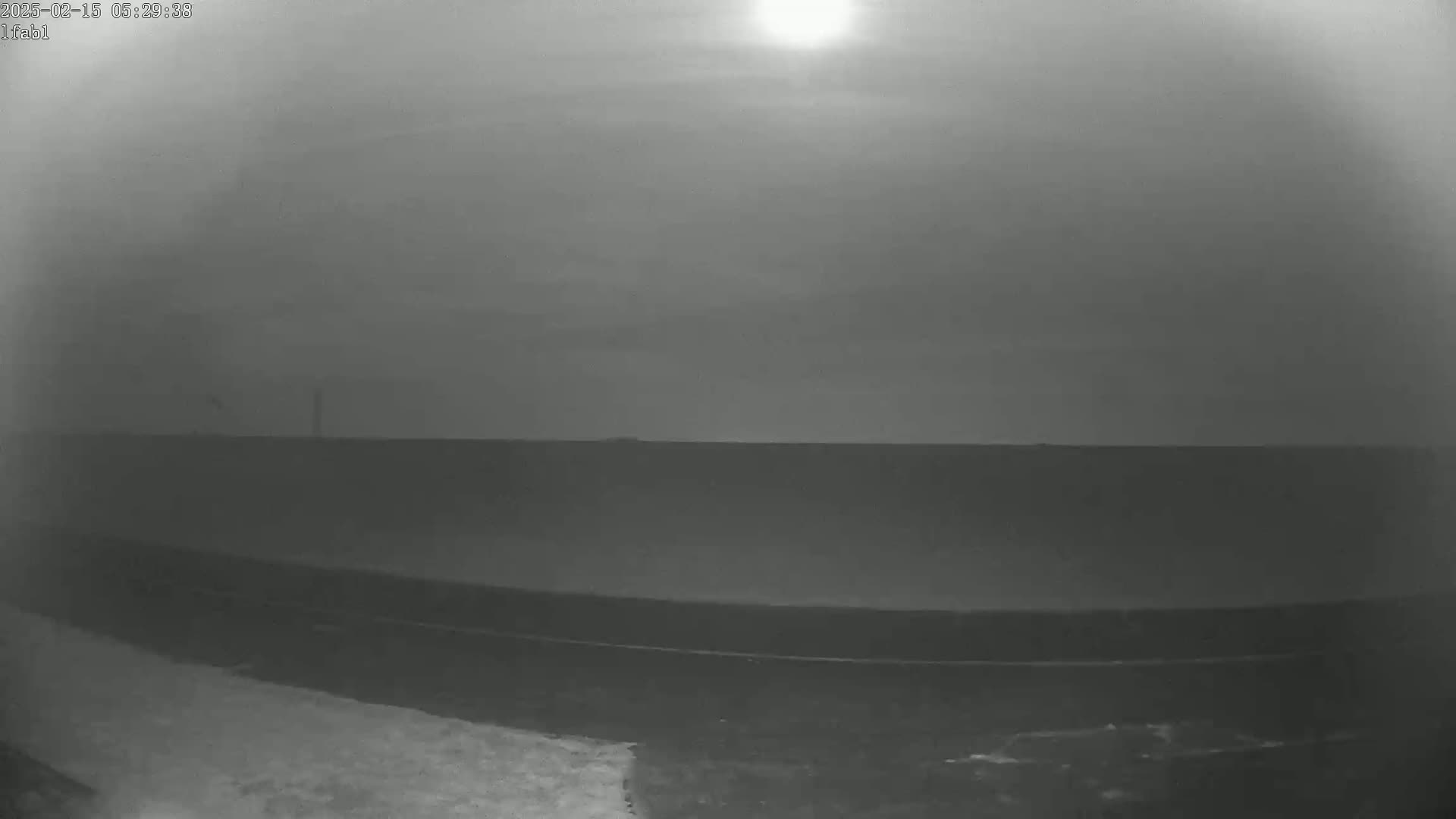 A hazy, bright sun shines over a flat, dark landscape under a mostly clear sky.