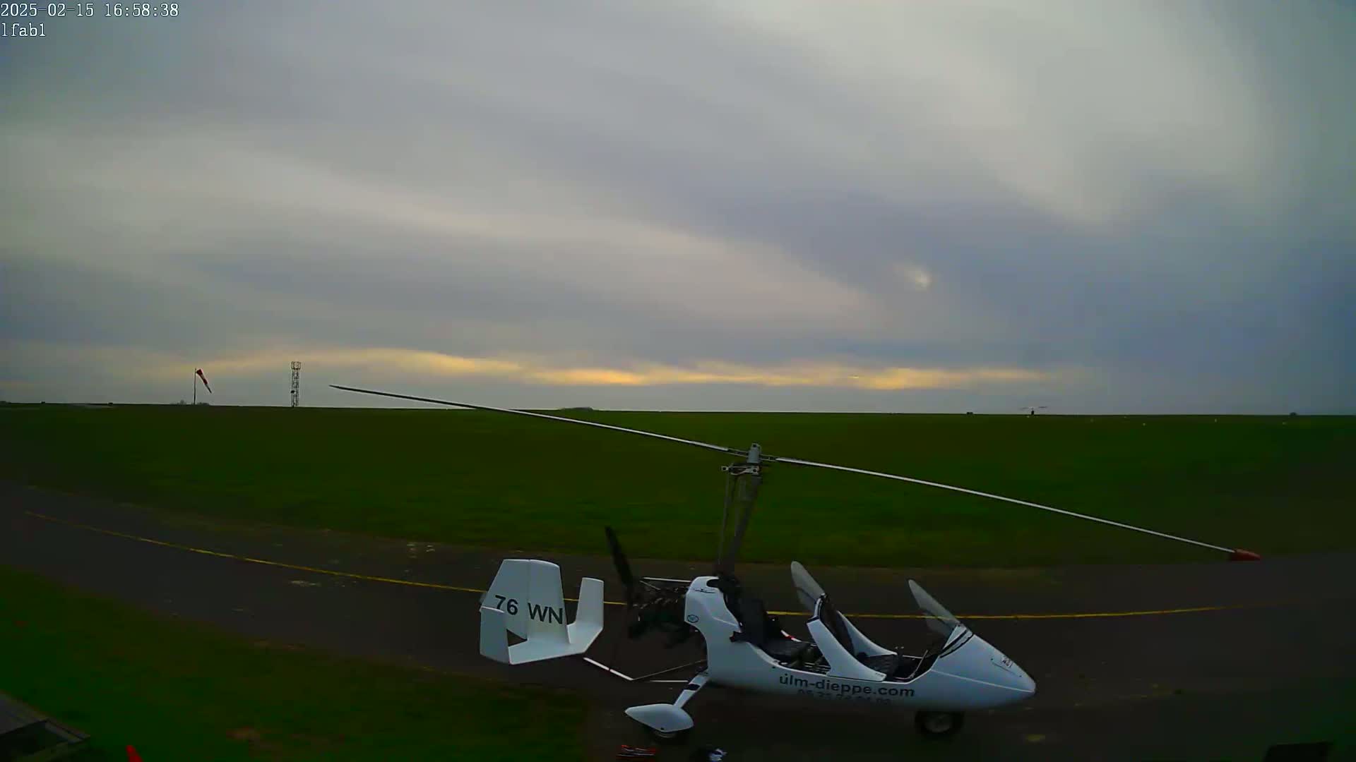 A white gyrocopter sits on a grassy airfield under an overcast sky.