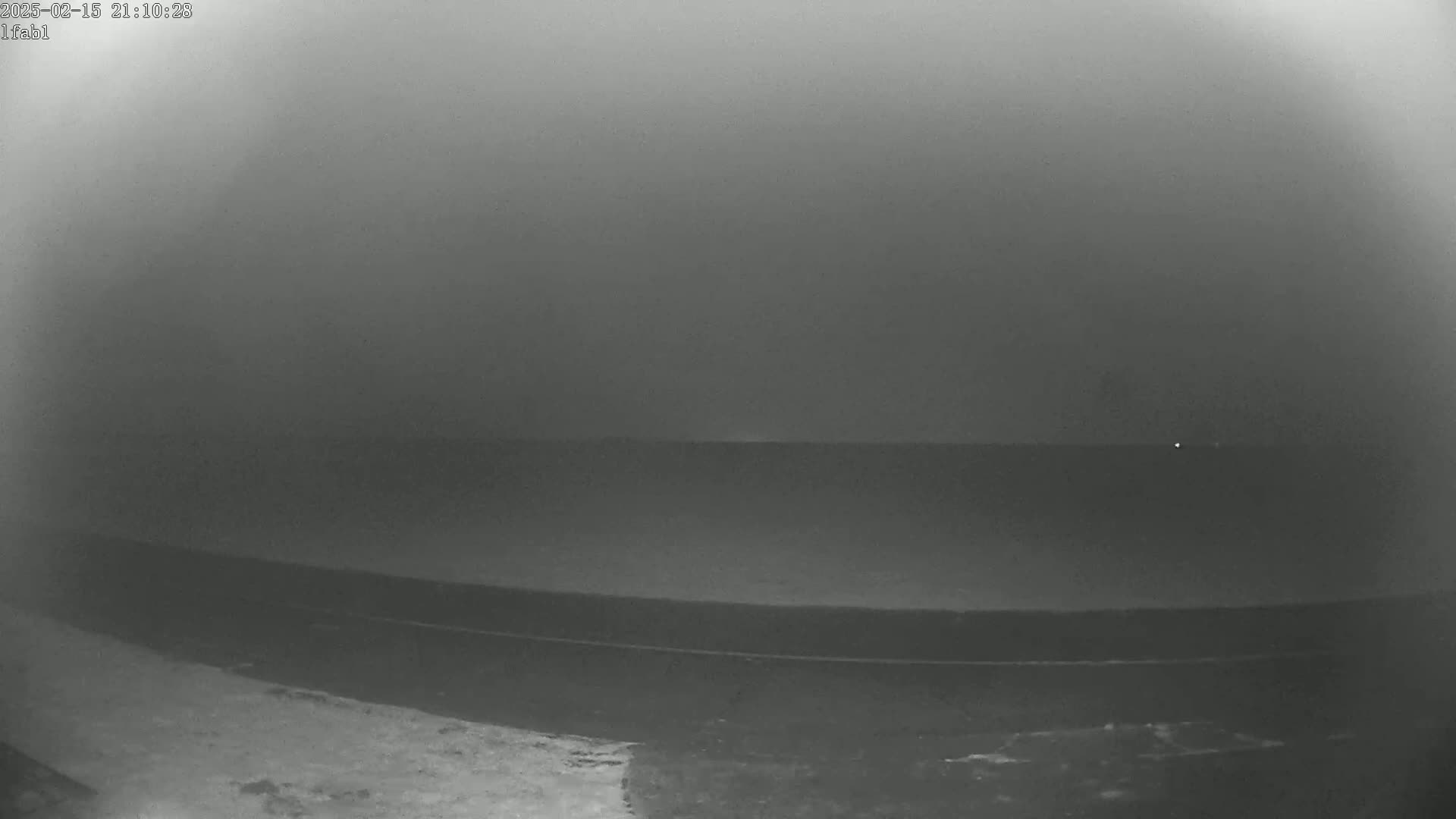A nighttime, foggy view shows a dark expanse of water and land, with a faint light visible in the distance.
