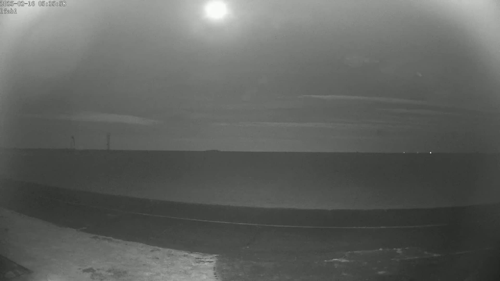 A bright sun illuminates a flat, dark landscape under a mostly clear, pale sky.