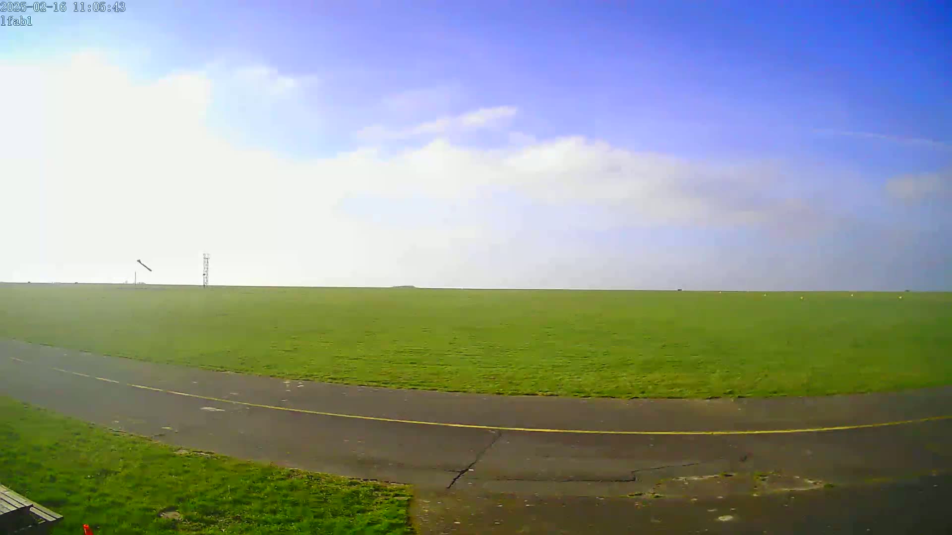 A paved path curves through a large, grassy field under a mostly clear, bright sky with a few scattered clouds.