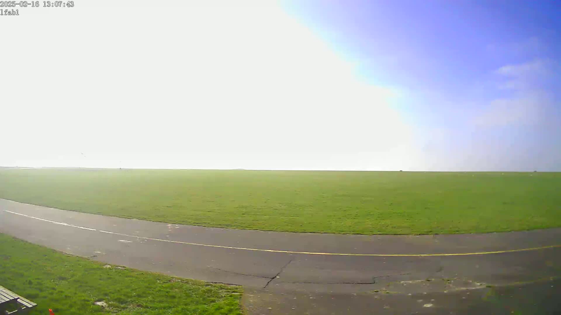 A paved path curves through a large, flat expanse of green grass under a mostly clear, bright sky.
