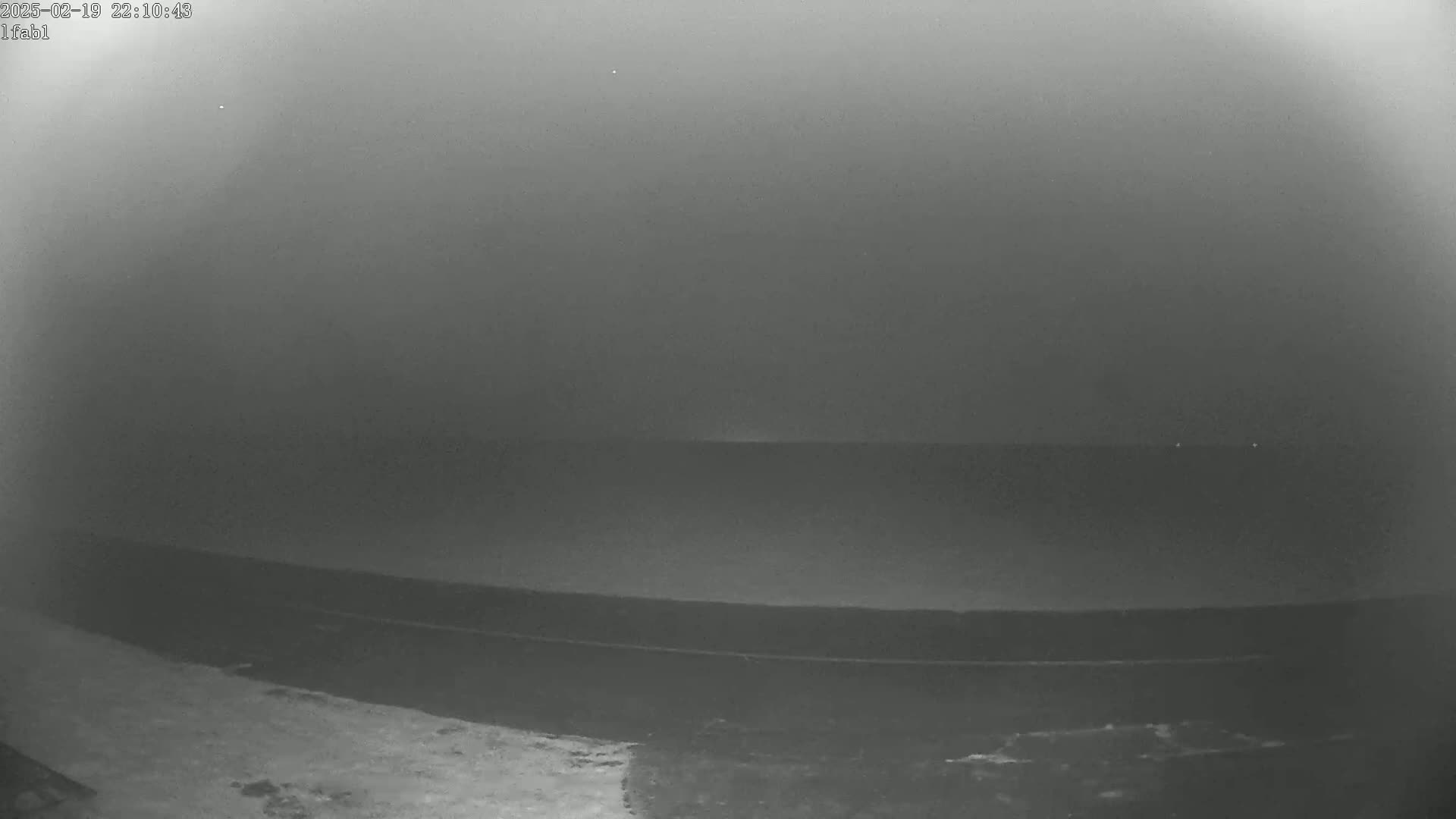 A nighttime, foggy view shows a dark body of water, possibly the ocean, with a dark shoreline in the foreground.