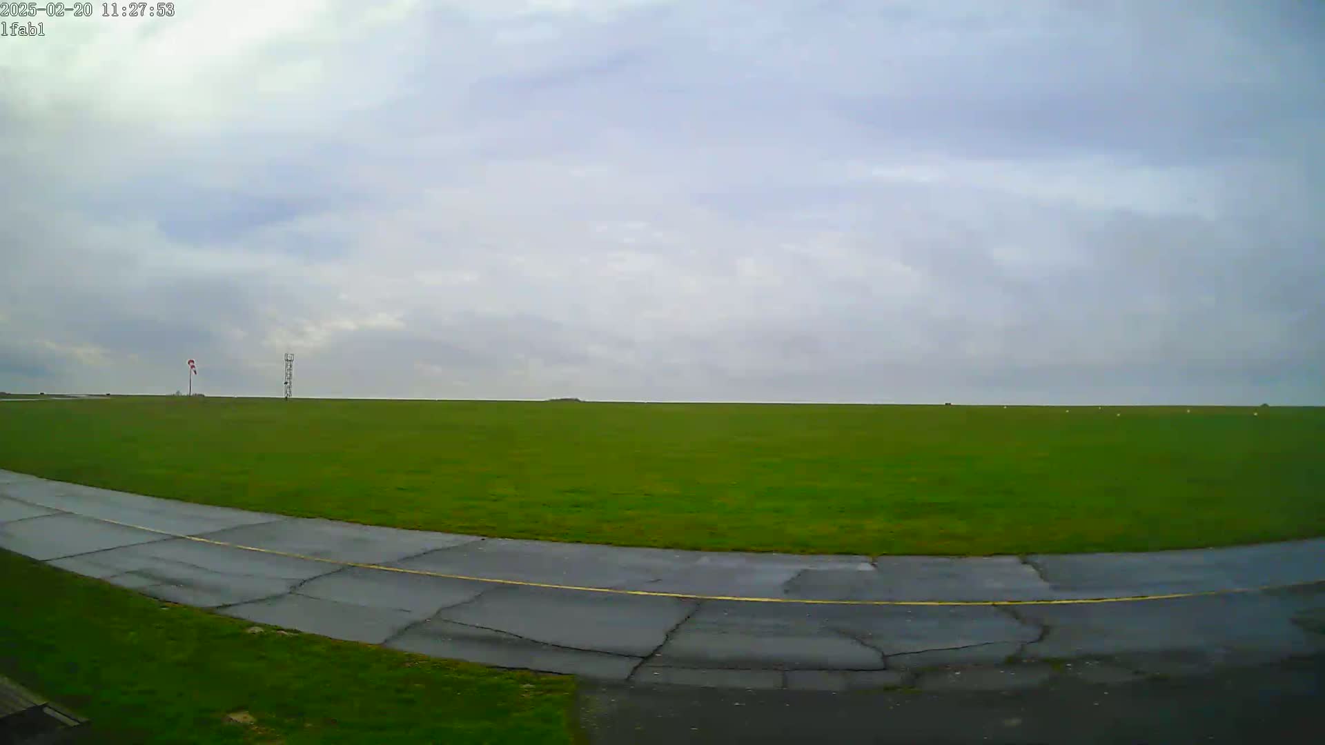 A paved taxiway curves through a large, grassy field under a mostly cloudy sky.