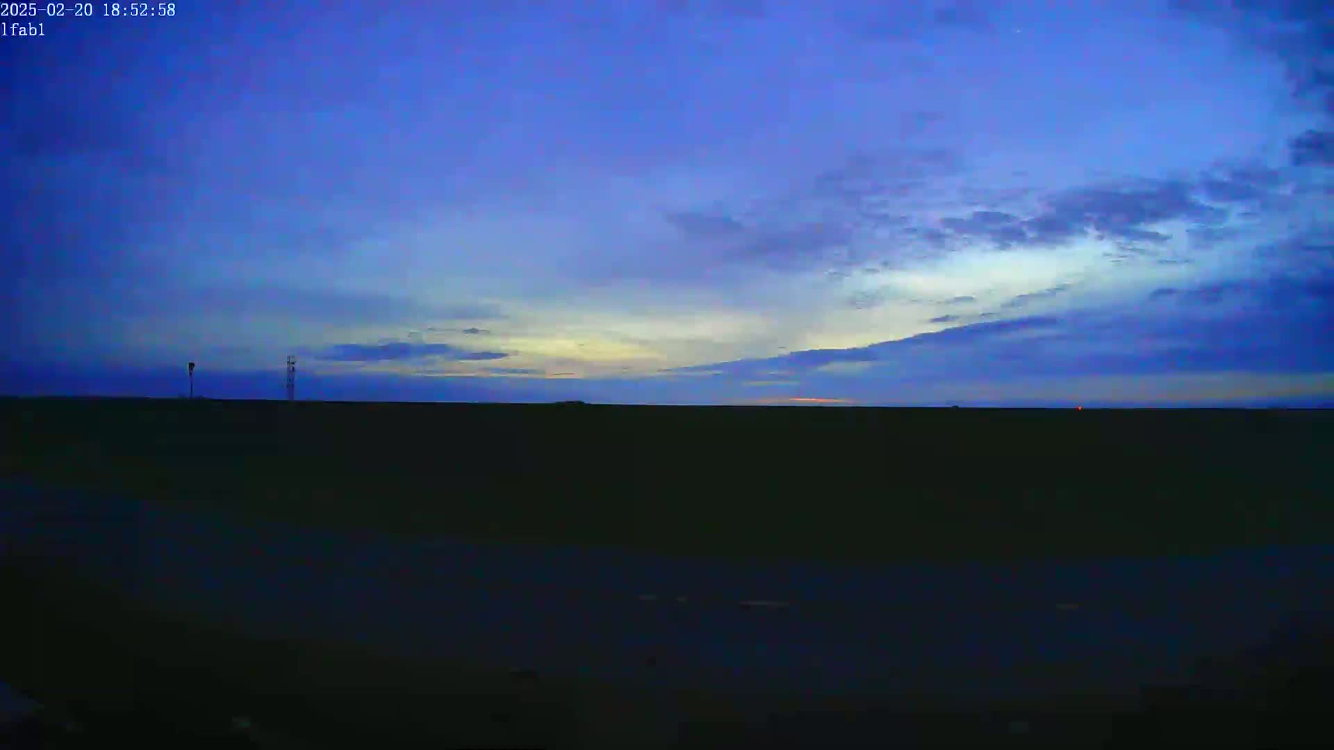 A twilight sky with thin clouds hangs over a dark, flat landscape featuring two distant, silhouetted towers.