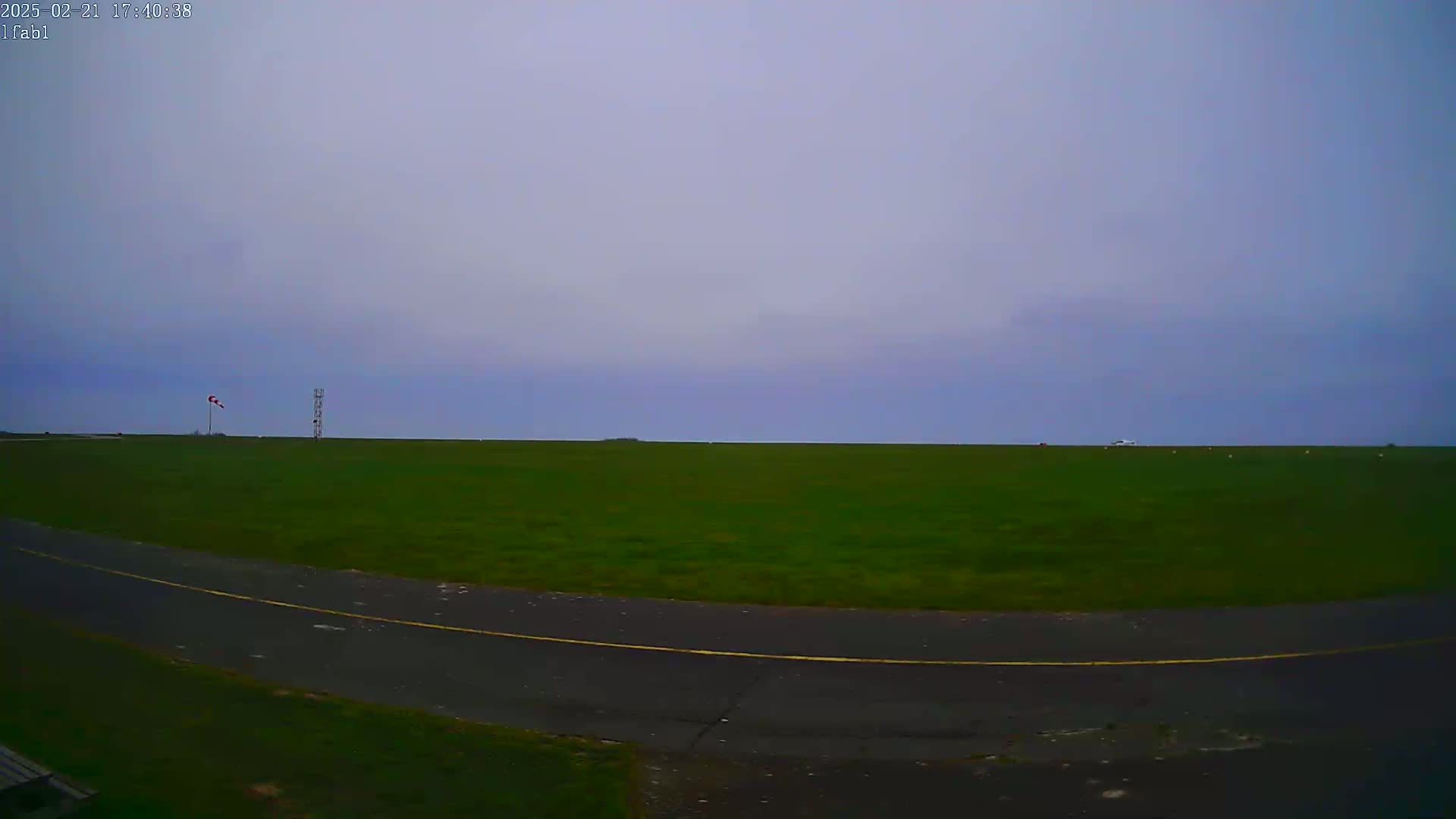 A vast, flat expanse of green grass is visible under an overcast sky, with a paved road curving in the foreground and a windsock and a tall structure in the mid-ground.