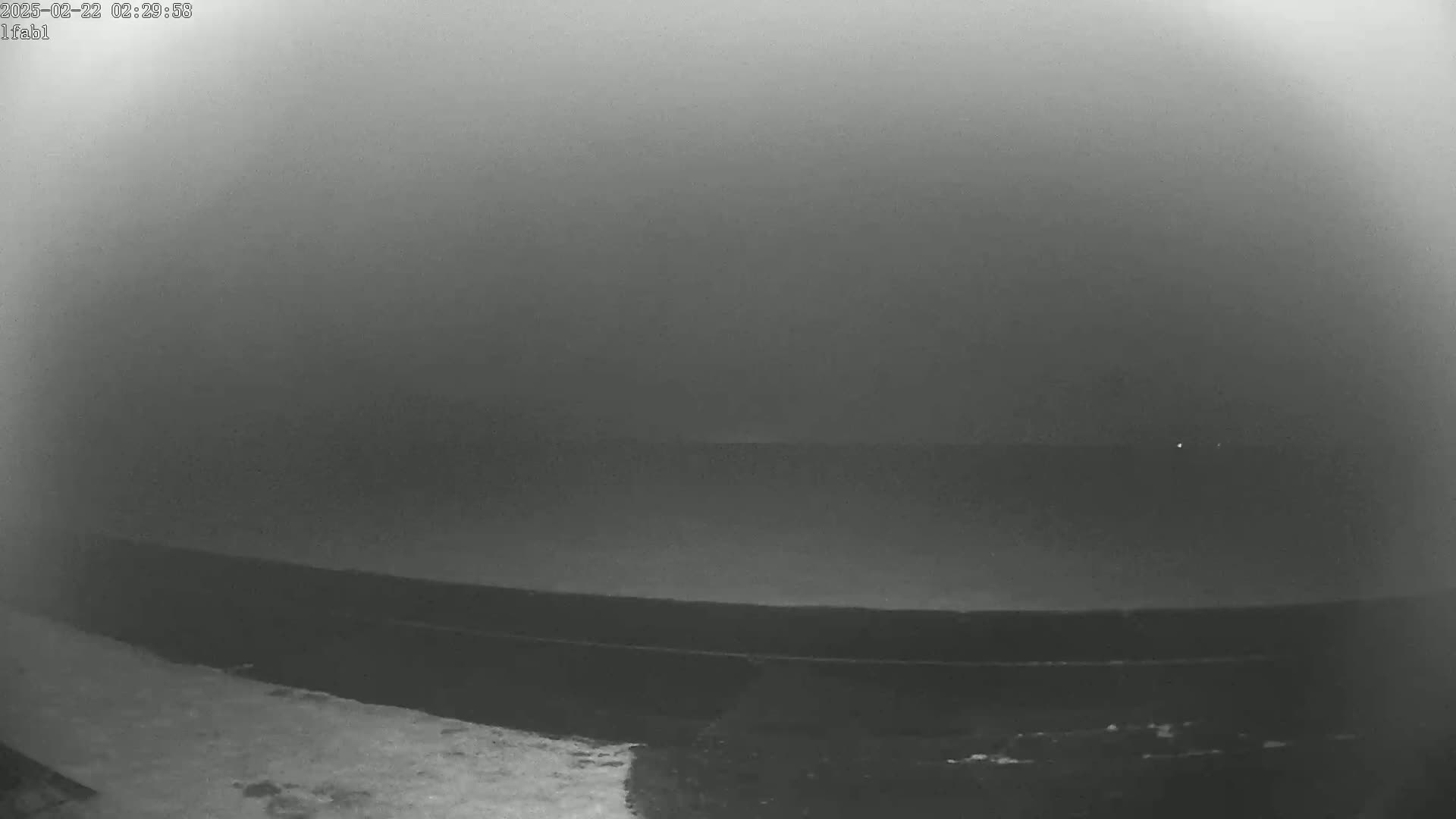 A low-visibility, foggy scene shows a dark expanse of water and land, possibly a shoreline, under a gray sky.