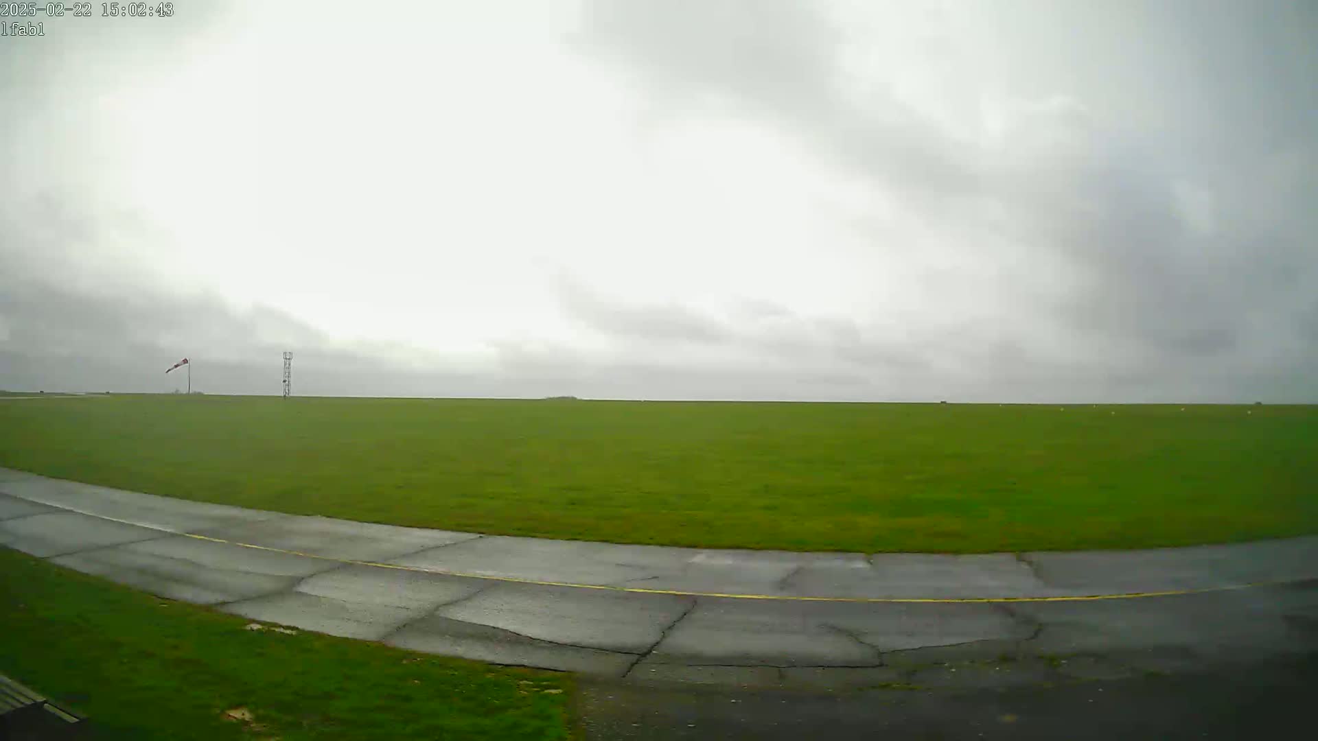 A wet, paved taxiway curves through a large, flat, grassy field under an overcast sky.