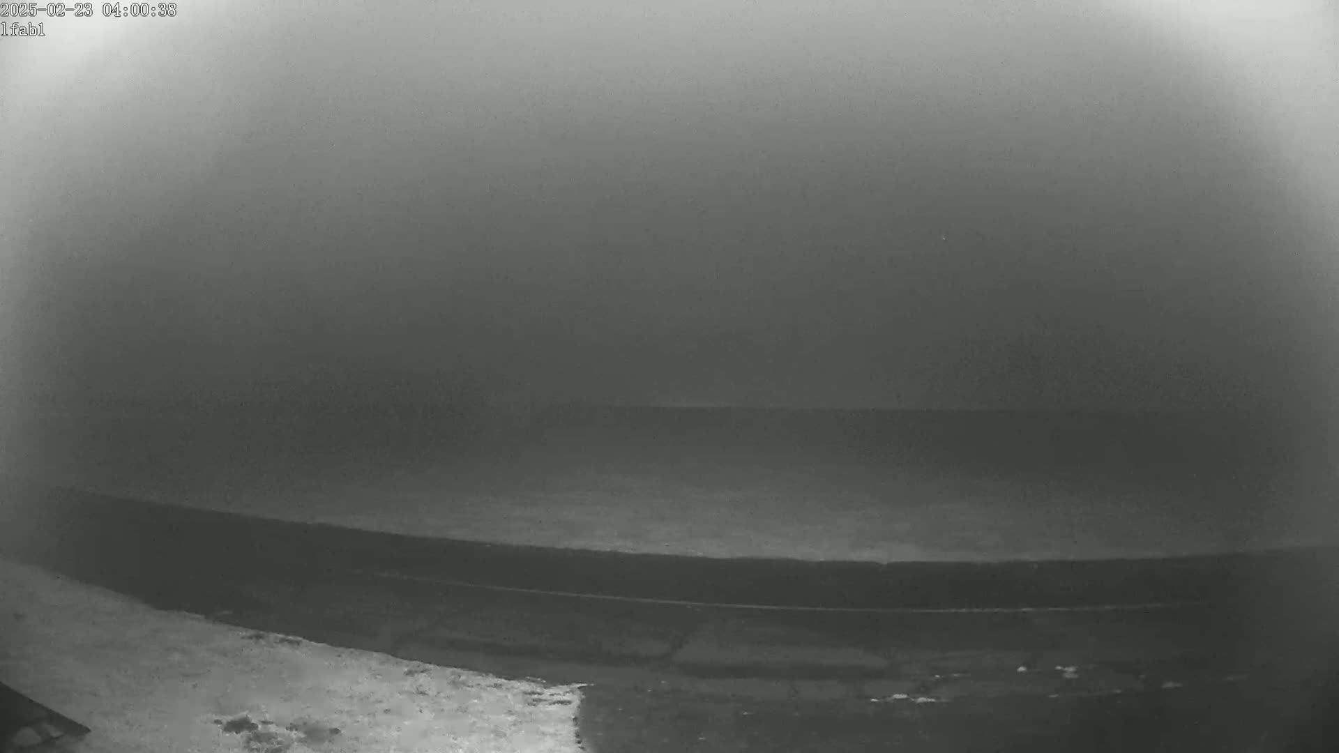 A low-visibility, foggy scene shows a dark, flat expanse of land or water near a lighter-colored edge.