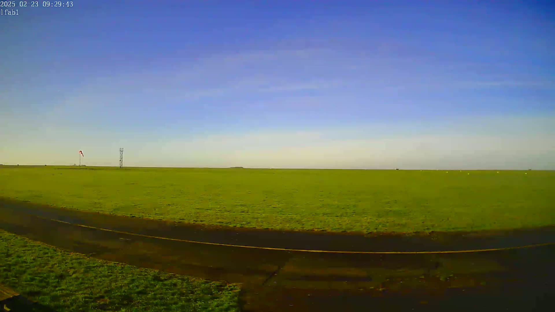 A vast, flat green field stretches to the horizon under a clear blue sky, with a paved road and a tall, thin structure visible in the middle ground.