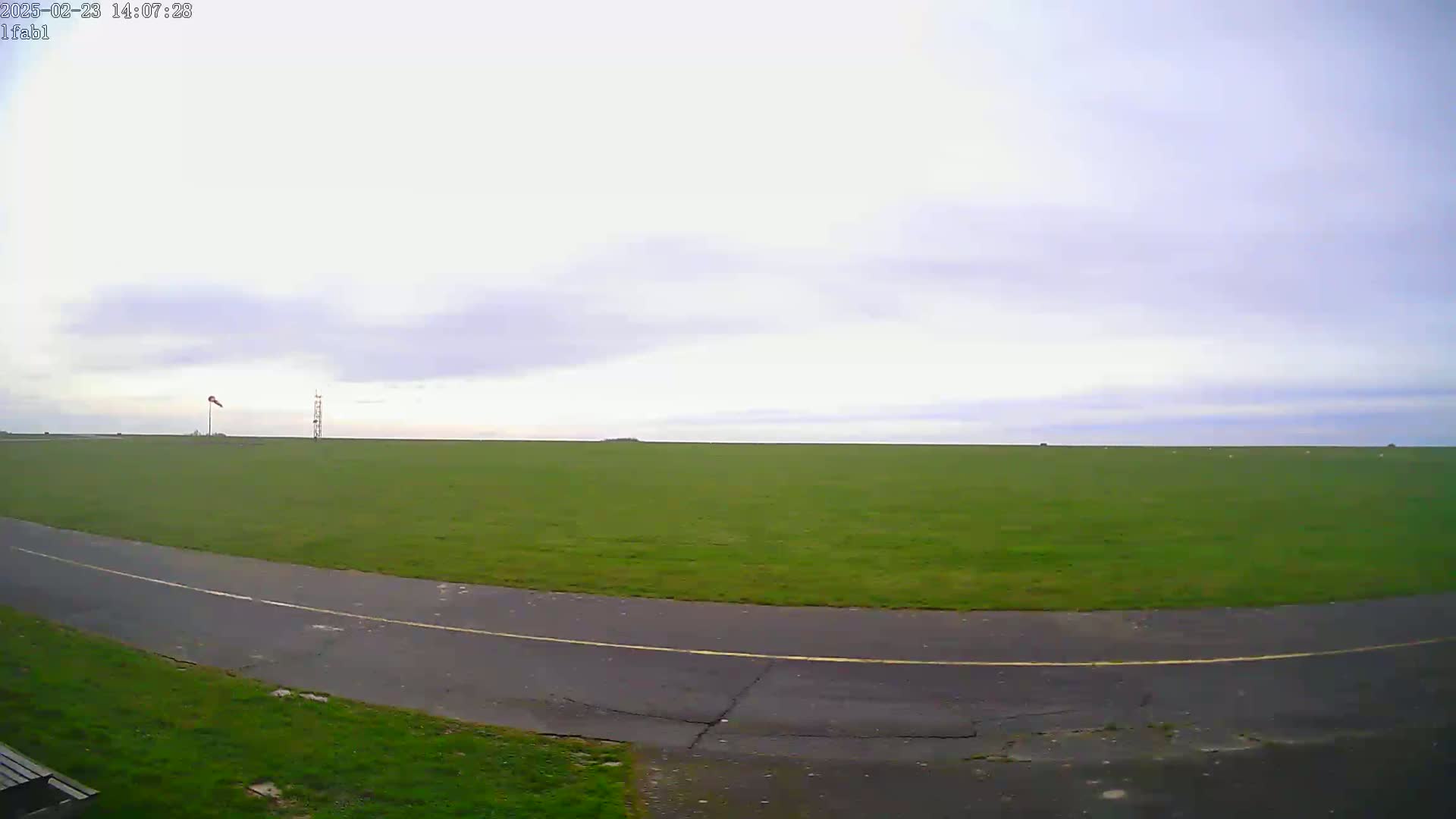 A paved road curves through a large, flat expanse of green grass under a mostly cloudy sky.