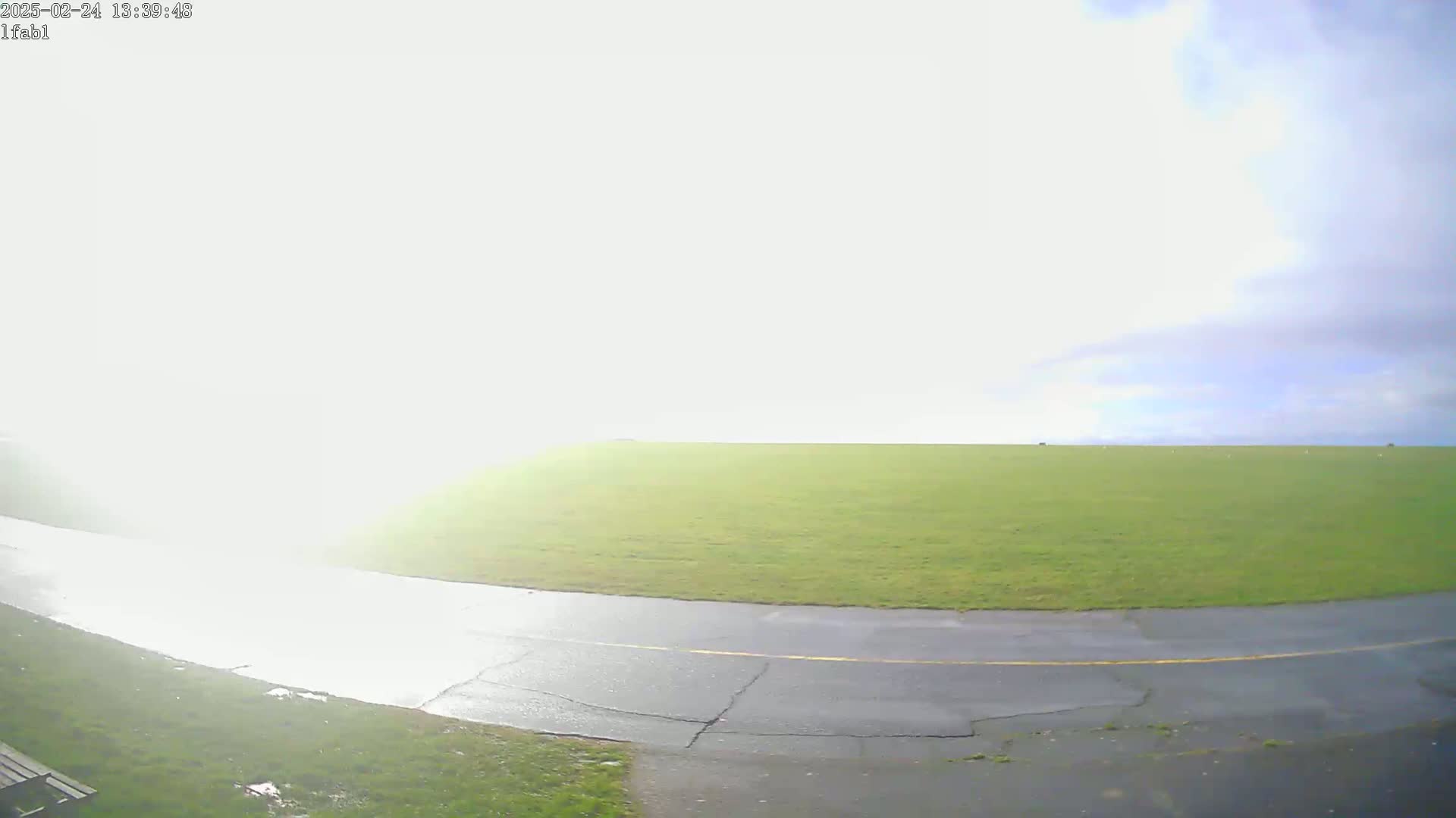 A paved road curves through a grassy field under a mostly cloudy sky, with some bright sunlight reflecting off the wet road.