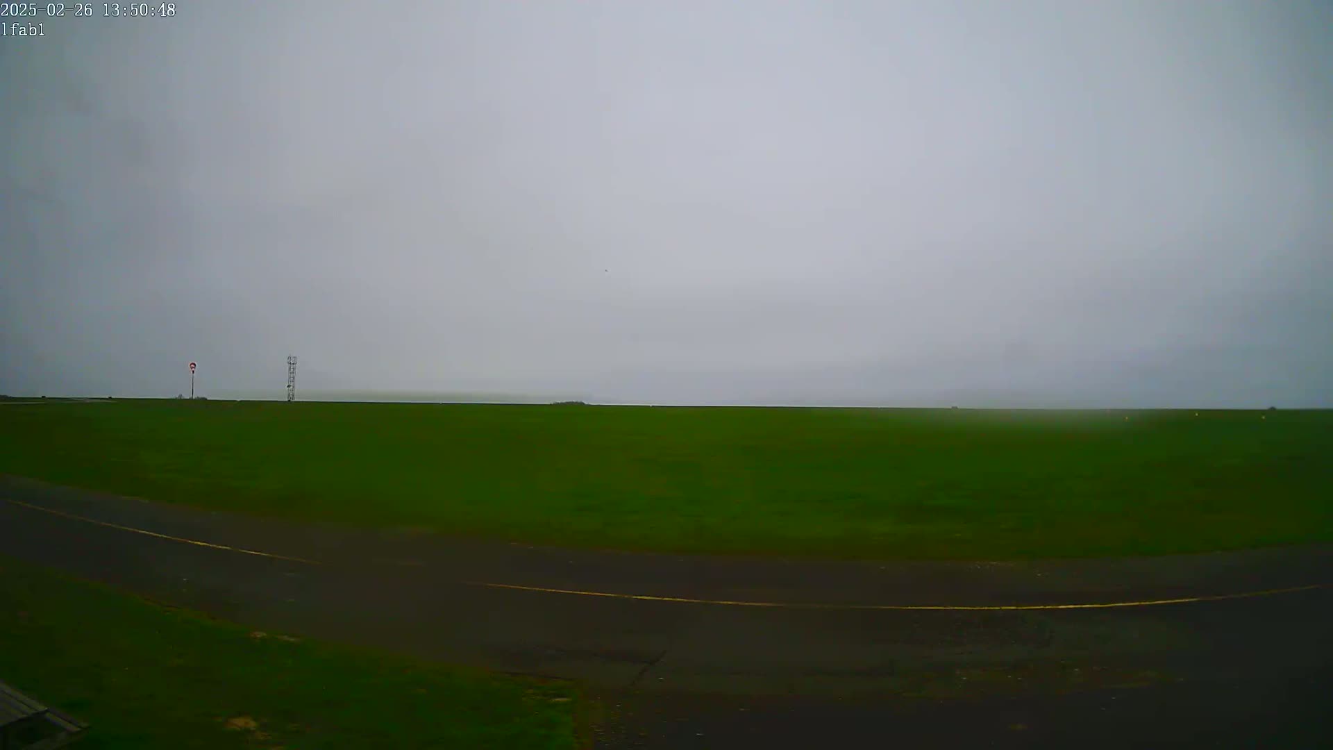 A flat, grassy field with a paved roadway curves into view under an overcast sky.