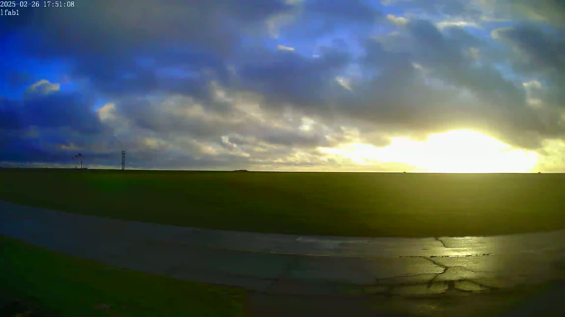 A wide, flat expanse of green land is visible, with a cracked asphalt road curving through the foreground, under a partly cloudy sky with a bright, low-hanging sun.