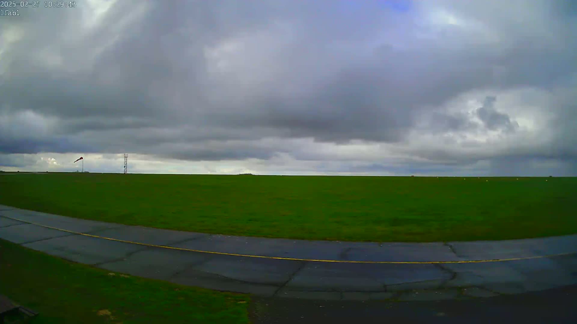 A wide, green grassy field stretches to the horizon under a cloudy sky, with a paved road curving in the foreground.