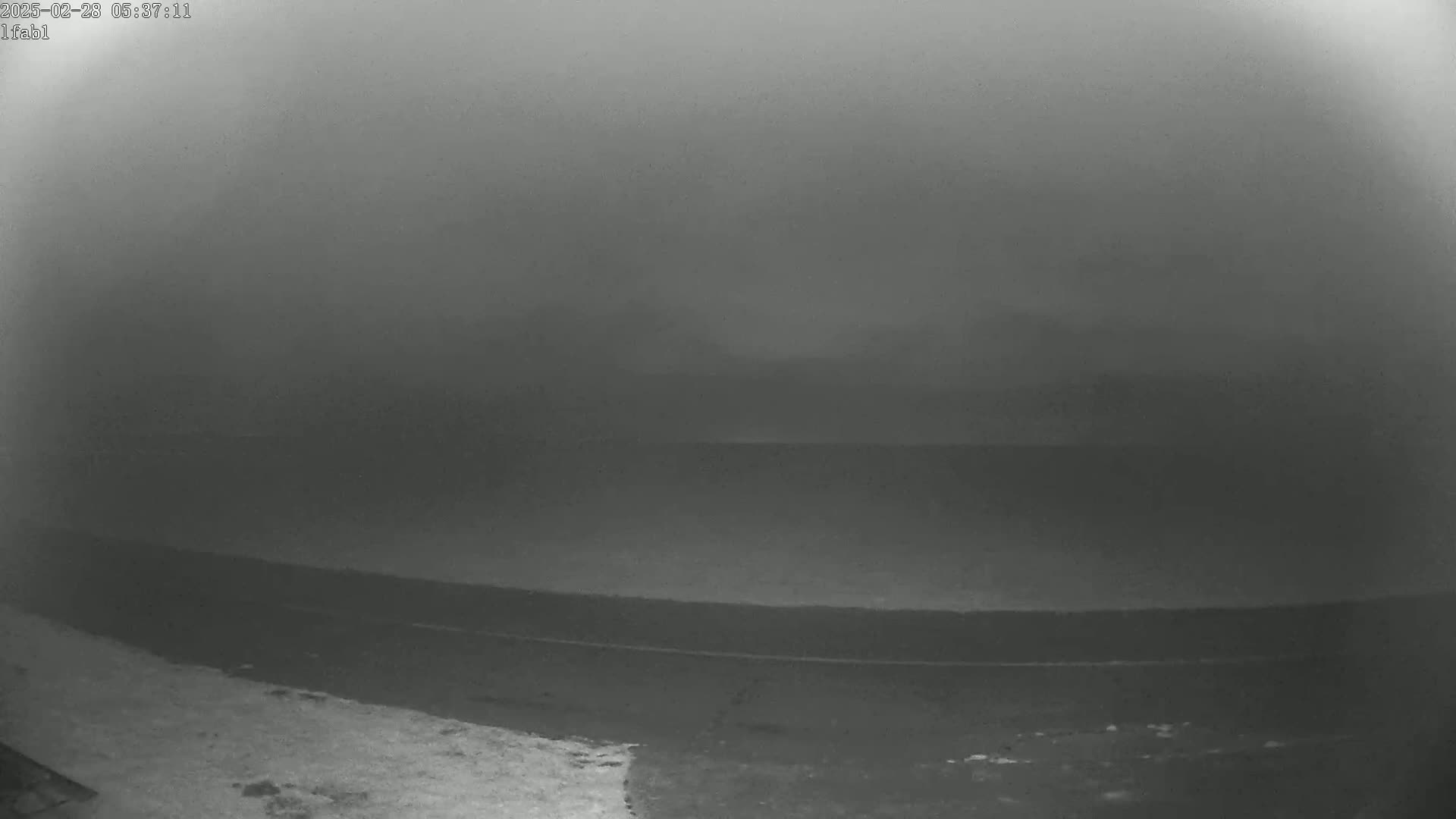A dark, foggy, or hazy landscape shows a flat expanse of land meeting a dark body of water under a muted sky.