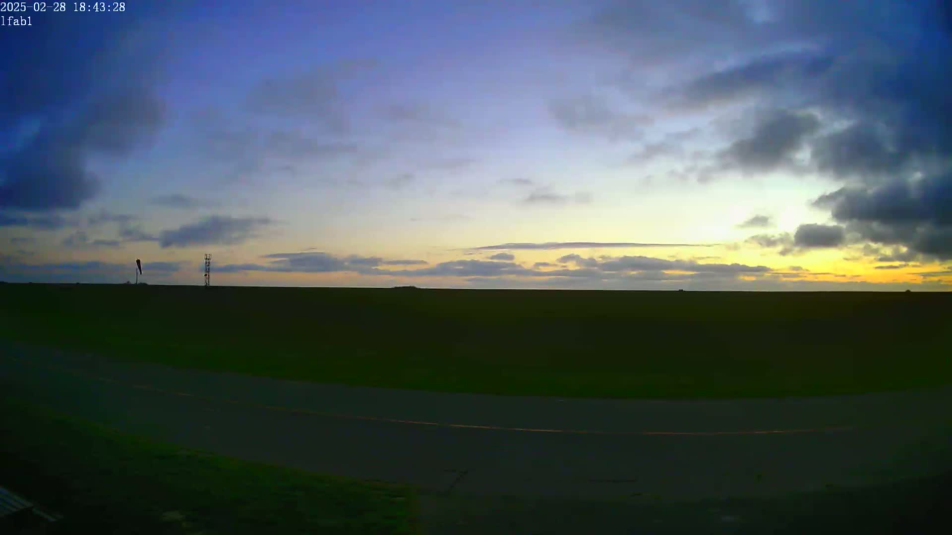 A flat, dark green field stretches to a pale yellow and blue sunset under a partly cloudy sky.