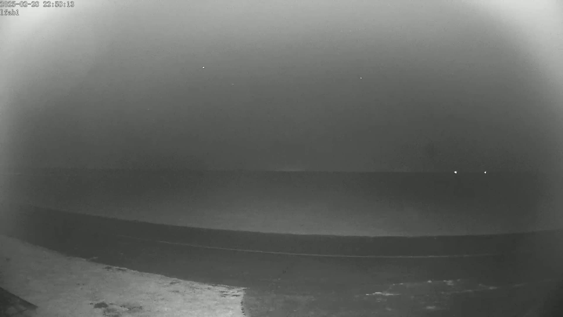 A dark, flat expanse of land or water is visible under a hazy, dark sky at night, with a few faint lights in the distance.