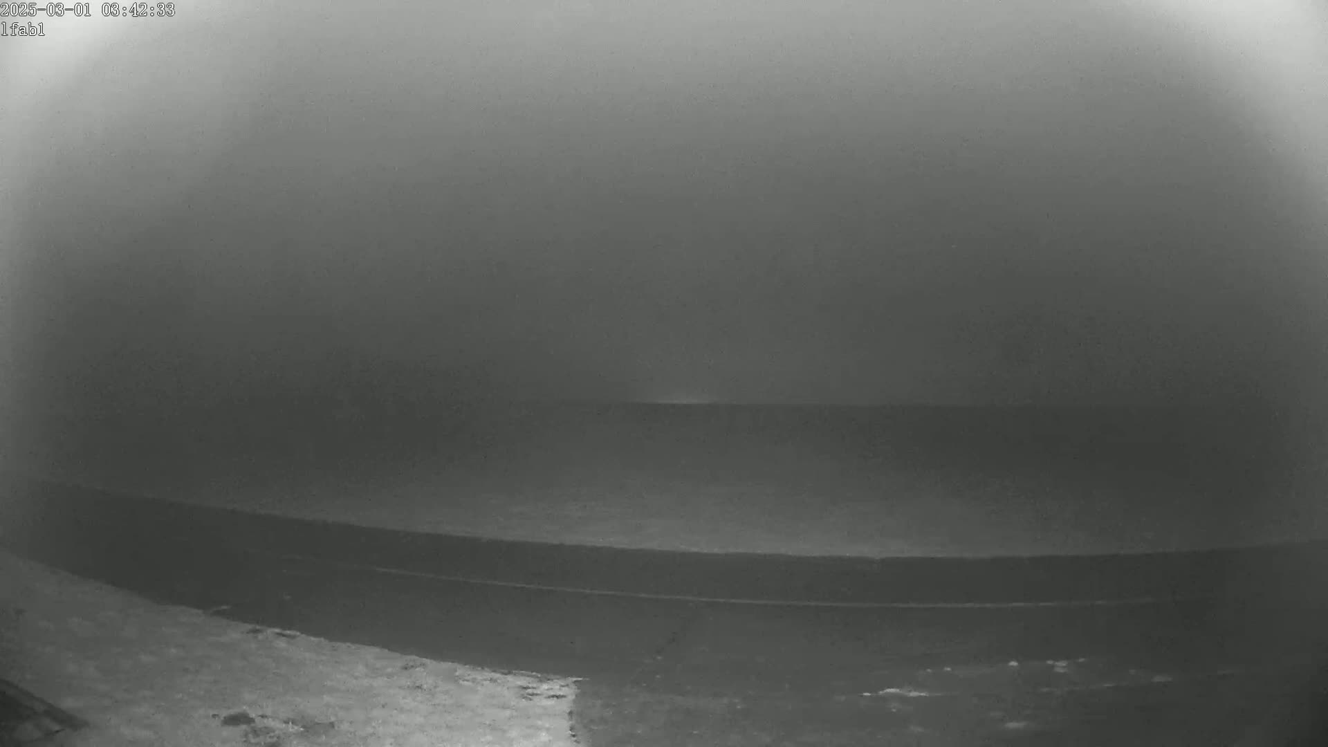A low-visibility, foggy scene shows a dark, flat expanse of land or water, possibly a road, extending into a thick fog.