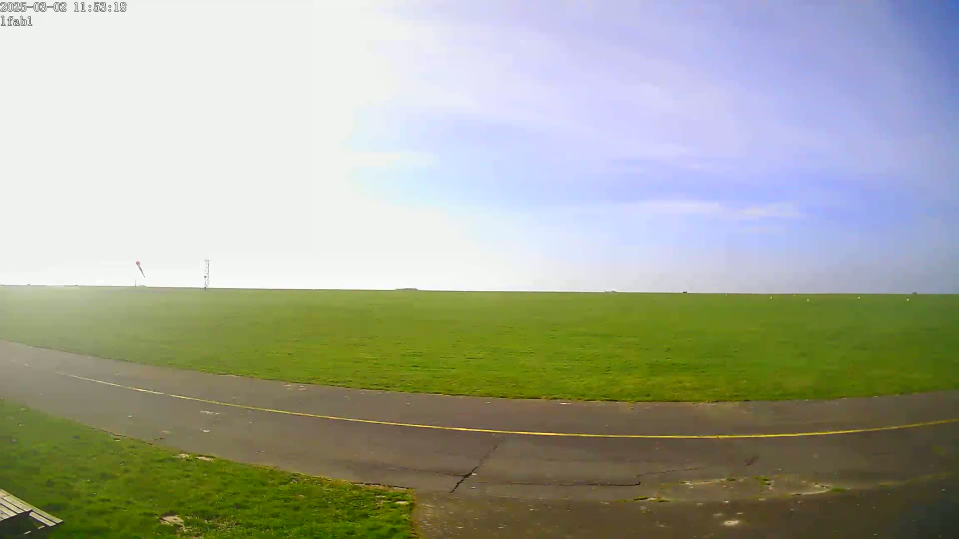 A paved path curves through a large, grassy field under a mostly clear, light blue sky.