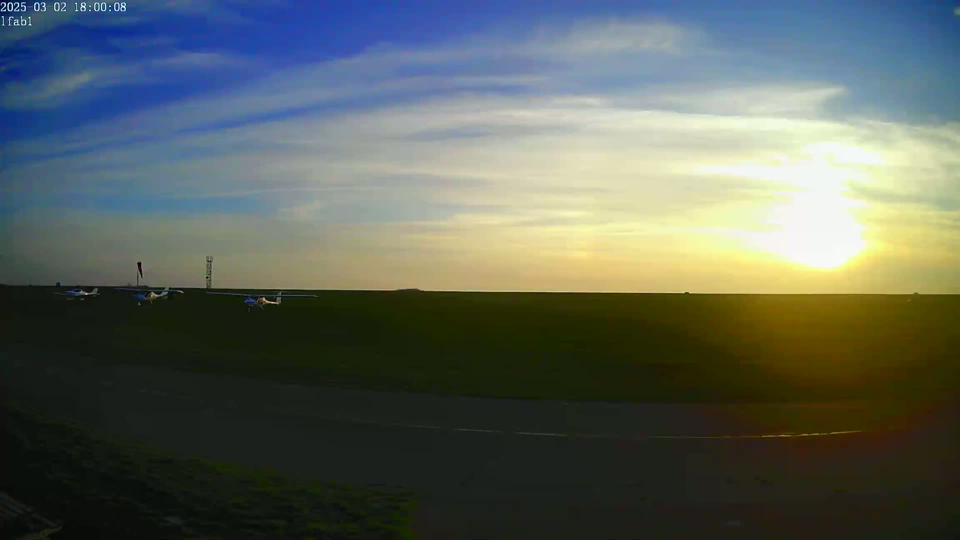 Several small airplanes are parked on a grassy airfield at sunset under a mostly clear sky.